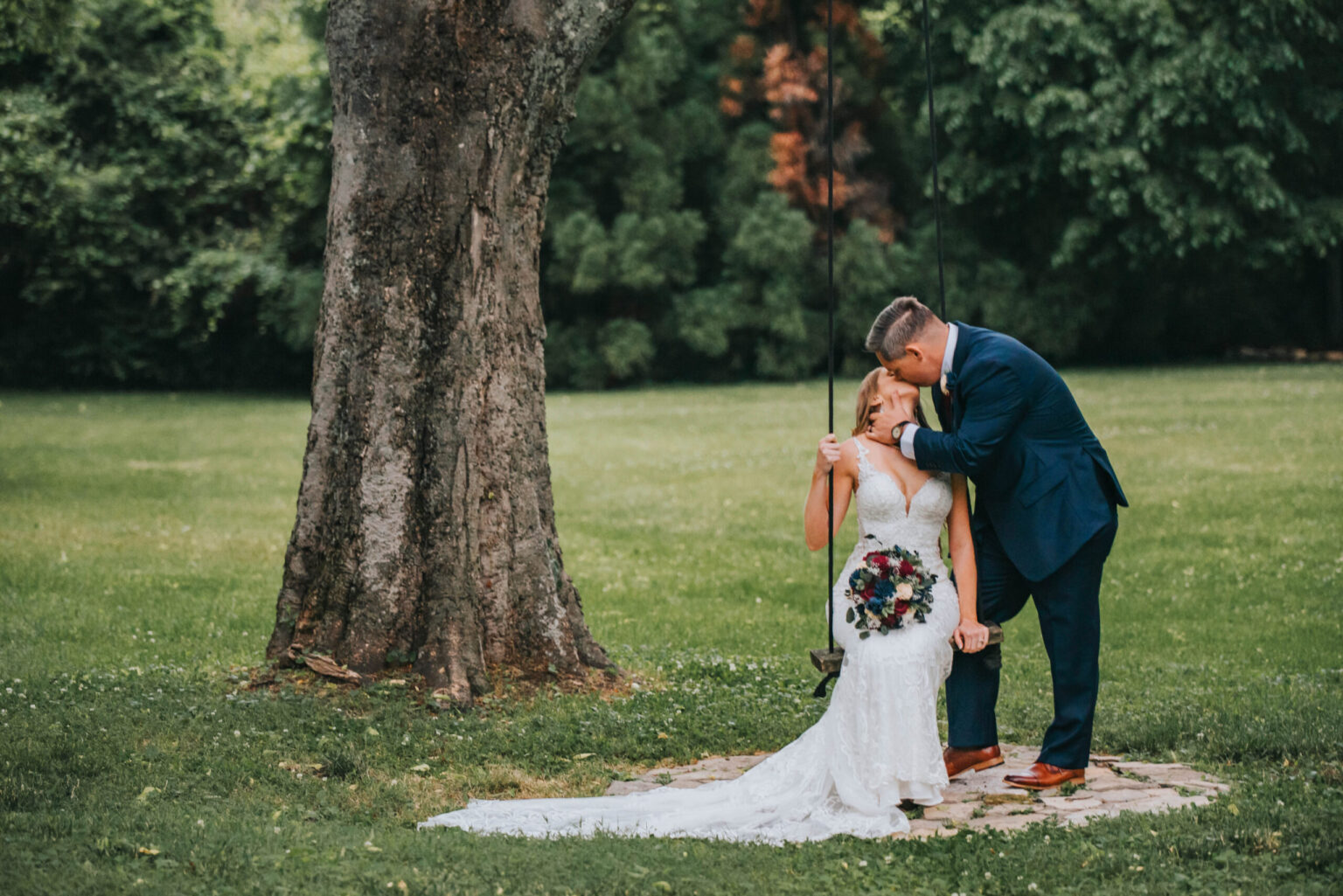 Elopement Wedding A bride in a white lace wedding dress holds a bouquet of purple flowers while sitting on a tree swing. A groom in a navy blue suit leans in to kiss her on the forehead. They are in a lush green park with dense trees in the background. Elopements Inc