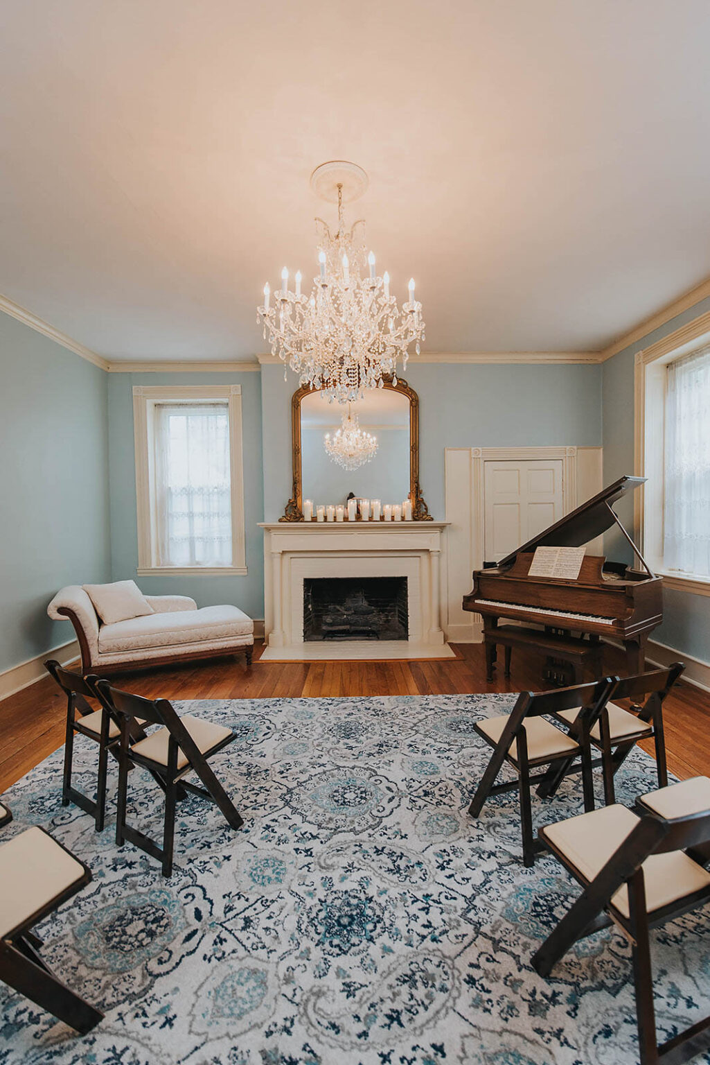 Elopement Wedding A room with light blue walls featuring a white fireplace, a grand piano, and a crystal chandelier. There is a white chaise lounge, several wooden folding chairs, and a rug with a blue and white pattern on the hardwood floor. A large mirror and candles sit on the mantle above the fireplace. Elopements Inc