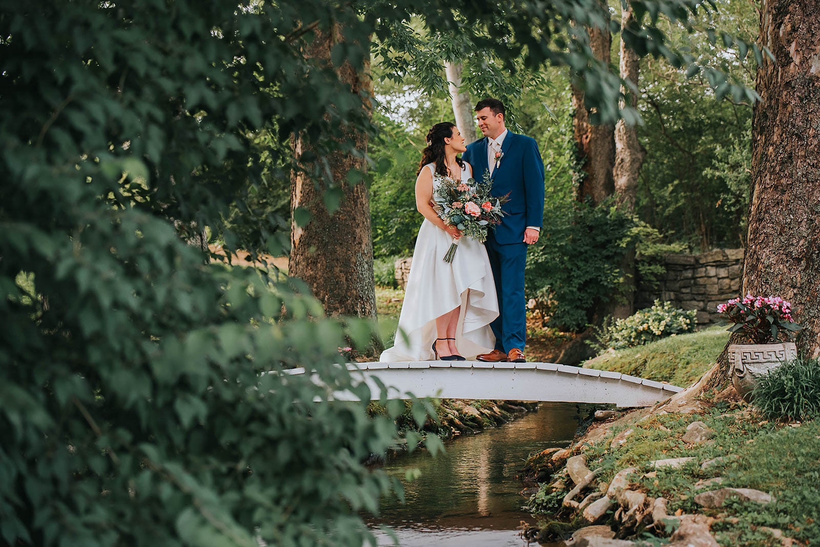 Elopement Wedding A bride and groom stand on a small white bridge over a stream in a lush, green garden. The bride is in a white wedding dress holding a bouquet of flowers, and the groom is in a blue suit. They are gazing at each other under the shade of large trees, with foliage surrounding them. Elopements Inc