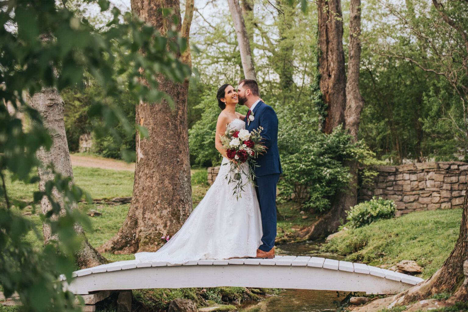 Elopement Wedding A bride in a white dress and groom in a blue suit stand closely on a small arched wooden bridge over a creek. They are surrounded by lush greenery and tall trees. The bride is holding a bouquet of red and white flowers, and the groom is kissing her on the cheek. Elopements Inc