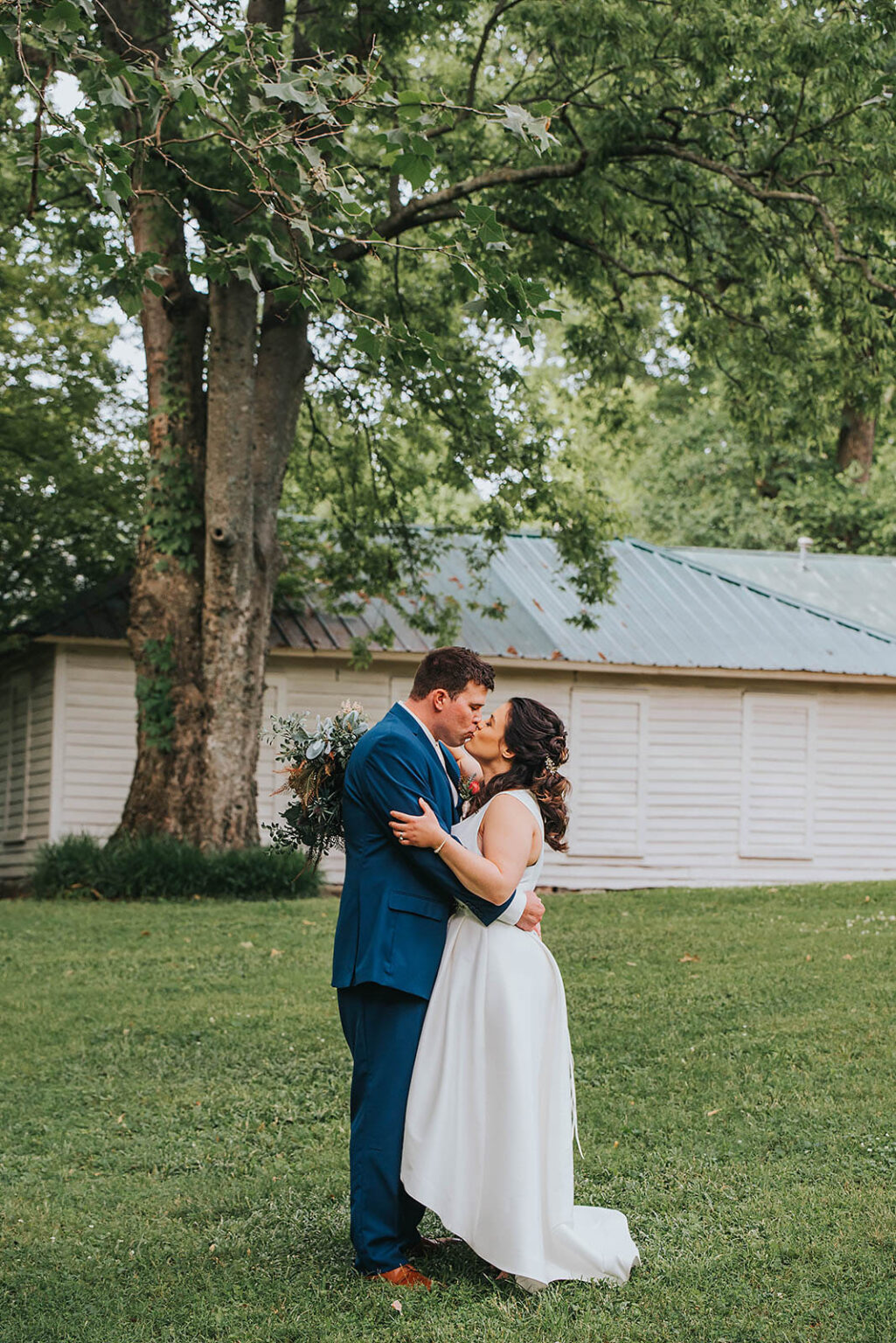 Elopement Wedding A bride and groom embrace and kiss in front of a white building with a metal roof and surrounded by lush green grass and trees. The bride wears a white dress, while the groom is dressed in a blue suit. The scene is outdoors, suggesting it might be a wedding or special event. Elopements Inc