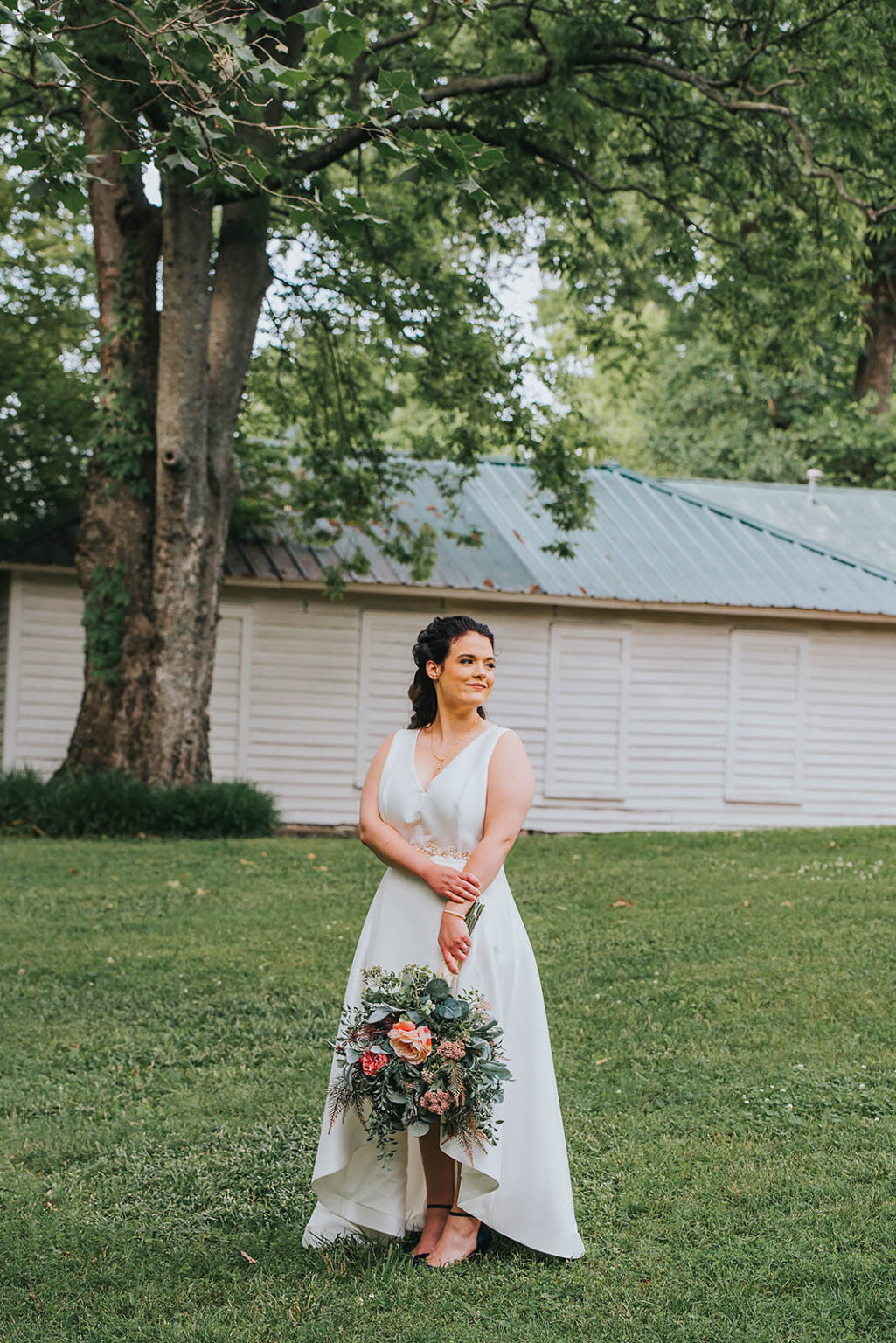 Elopement Wedding A woman wearing a sleeveless white dress stands barefoot on a lawn, holding a bouquet of flowers. She has dark hair styled in an updo and is adorned with a necklace and bracelet. Behind her, there is a large tree and a white building with a green metal roof, surrounded by lush greenery. Elopements Inc