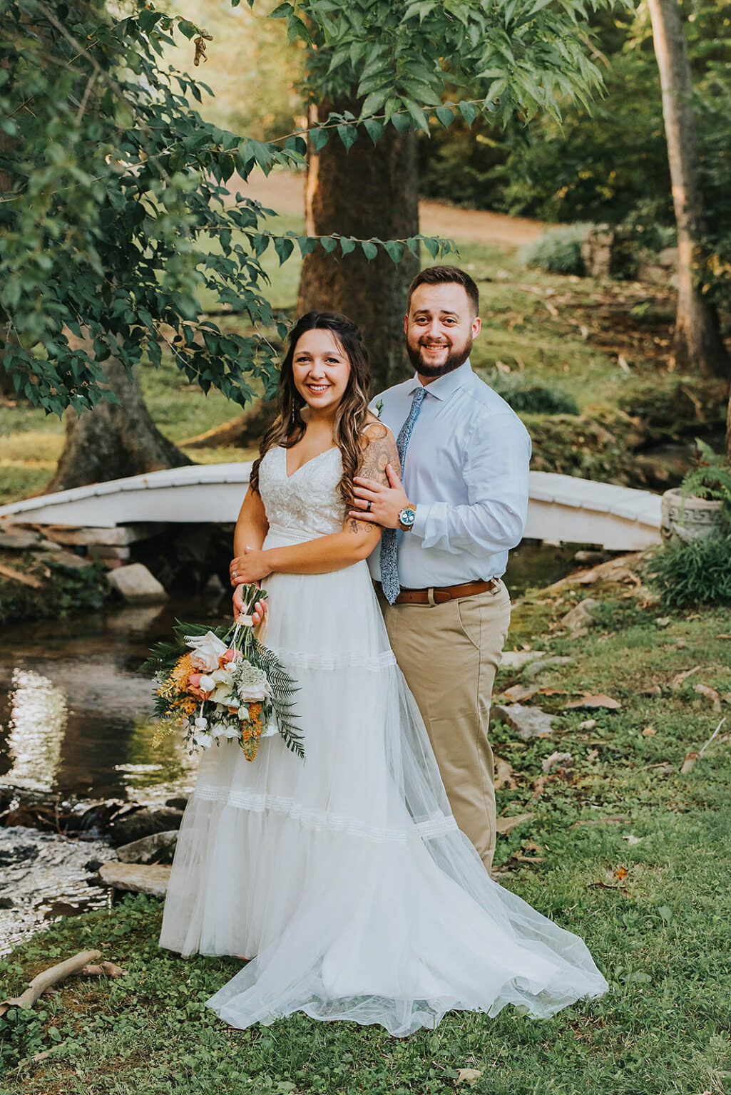 Elopement Wedding A bride in a white dress and groom in a light blue shirt and beige pants stand smiling by a small stream with a white wooden bridge in the background. The bride holds a bouquet of flowers as the groom wraps his arm around her waist. Both appear happy, surrounded by greenery and trees. Elopements Inc