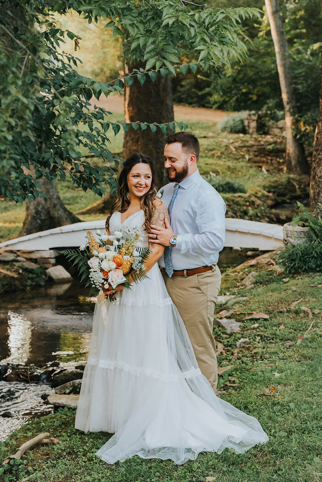 Elopement Wedding A smiling bride in a white dress holds a bouquet while a groom in a light blue shirt and khaki pants stands behind her, touching her shoulder and waist. They stand beside a small stream, with a white arched bridge and lush greenery in the background. It’s a sunny outdoor setting. Elopements Inc