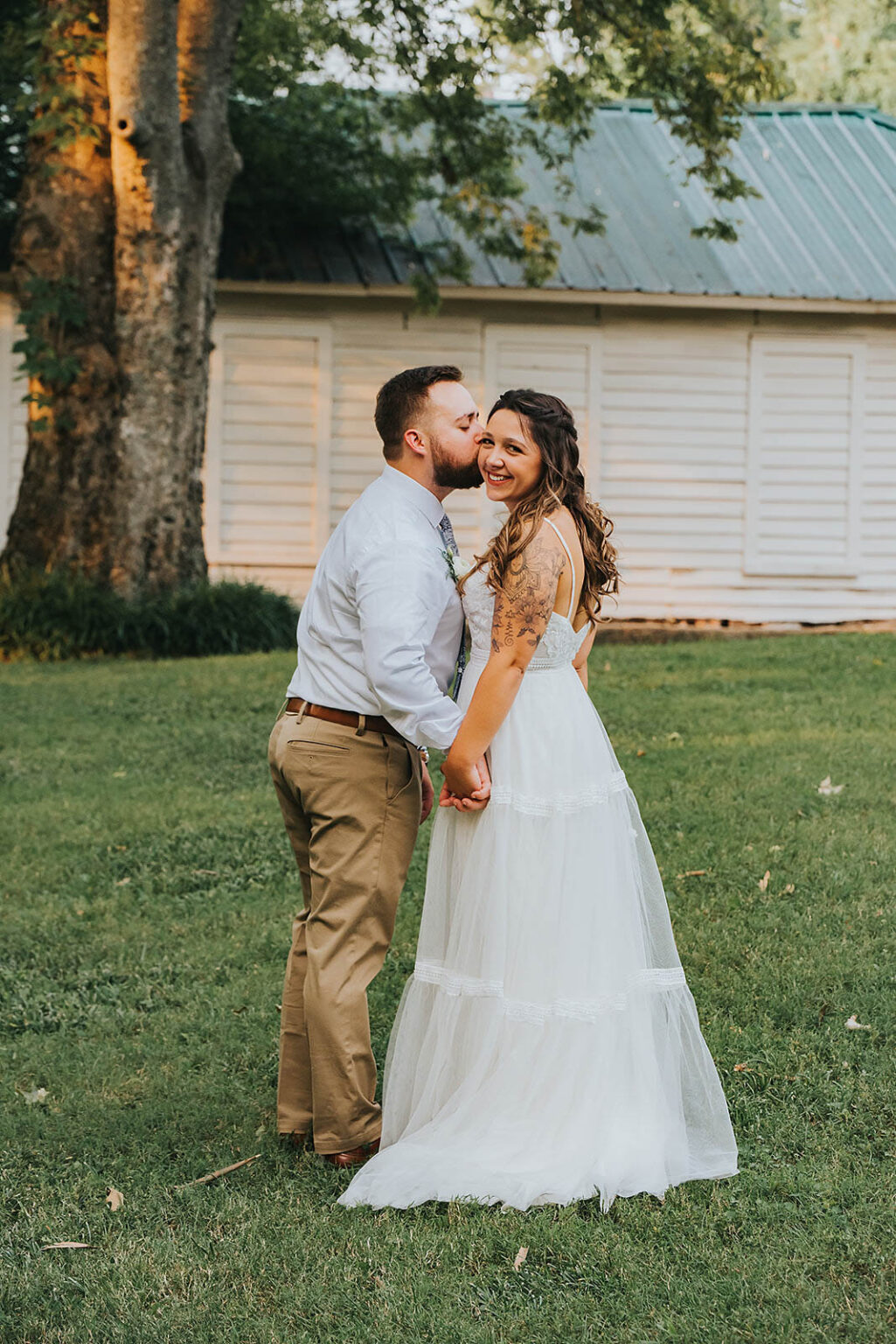 Elopement Wedding A newlywed couple stands outdoors on a grassy lawn. The bride, in a flowing white gown, smiles at the camera while the groom, dressed in a light shirt and khaki pants, kisses her on the temple. They hold hands, with a wooden building and trees in the background. Elopements Inc