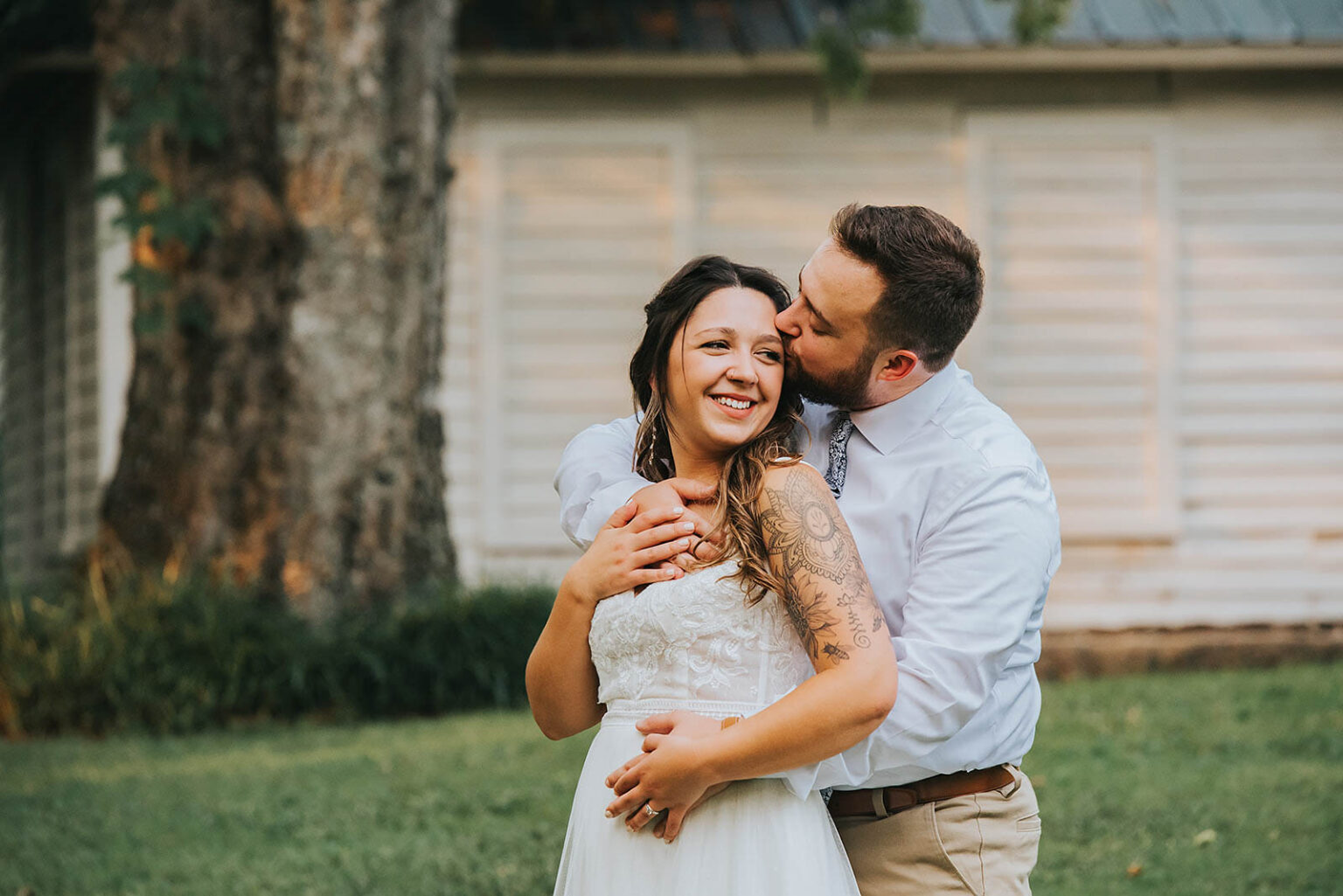 Elopement Wedding A couple stands outside in front of a white house with green shutters. The man, wearing a white shirt and beige pants, embraces the woman from behind and kisses her on the cheek. The woman, in a white sleeveless dress, smiles joyfully. They stand on green grass with trees in the background. Elopements Inc