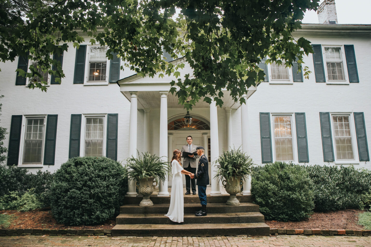 Elopement Wedding A couple is getting married on the front steps of a large white house with black shutters and large columns. They face each other while holding hands, and an officiant stands between them. Green shrubbery surrounds the scene, and a large tree provides a canopy overhead. Elopements Inc