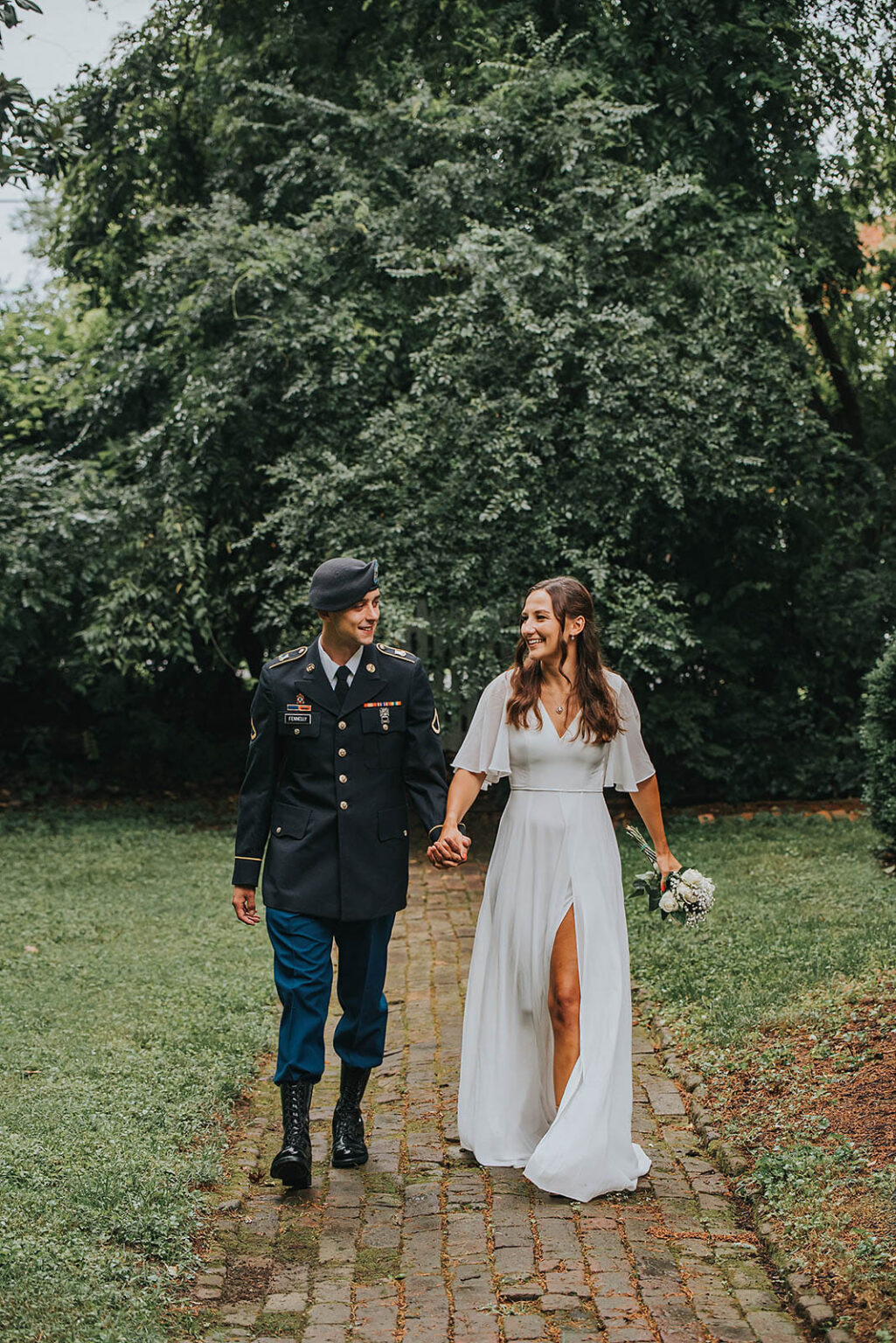 Elopement Wedding A man in a military uniform and a woman in a white wedding dress walk hand-in-hand along a cobblestone path, surrounded by lush greenery. The man wears a beret and boots, while the woman holds a small bouquet and smiles at him. Their expressions suggest happiness as they walk together. Elopements Inc