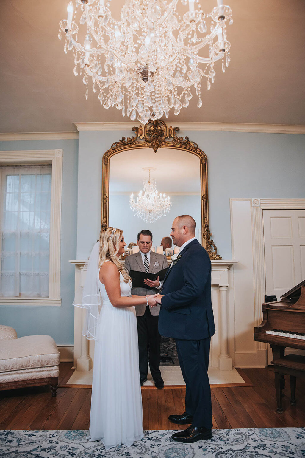 Elopement Wedding A bride and groom stand facing each other, holding hands in front of a fireplace with an ornate mirror and a chandelier above. The bride wears a white gown and veil, the groom a dark suit. Behind them, an officiant conducts the ceremony. The room is elegantly decorated with light blue walls and classical furnishings. Elopements Inc