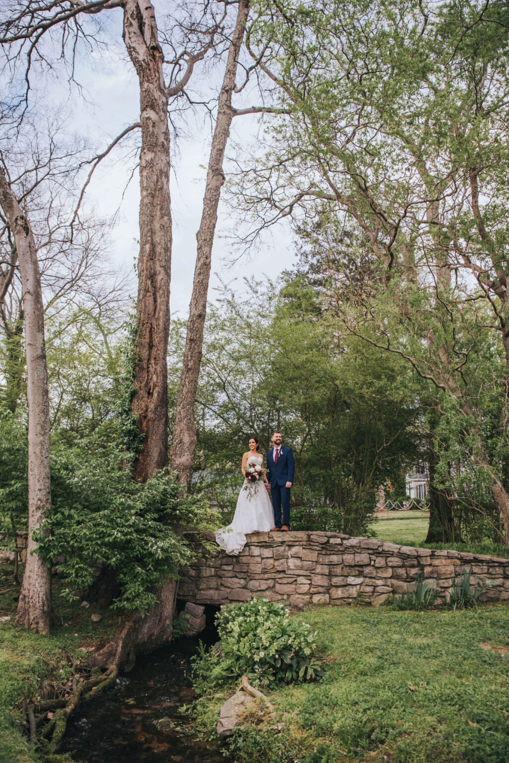 Elopement Wedding A bride in a white gown and a groom in a navy suit stand together on a small stone bridge in a lush, green woodland area. Tall, slender trees surround them, and a narrow stream flows beneath the bridge, creating a peaceful, romantic scene. Elopements Inc