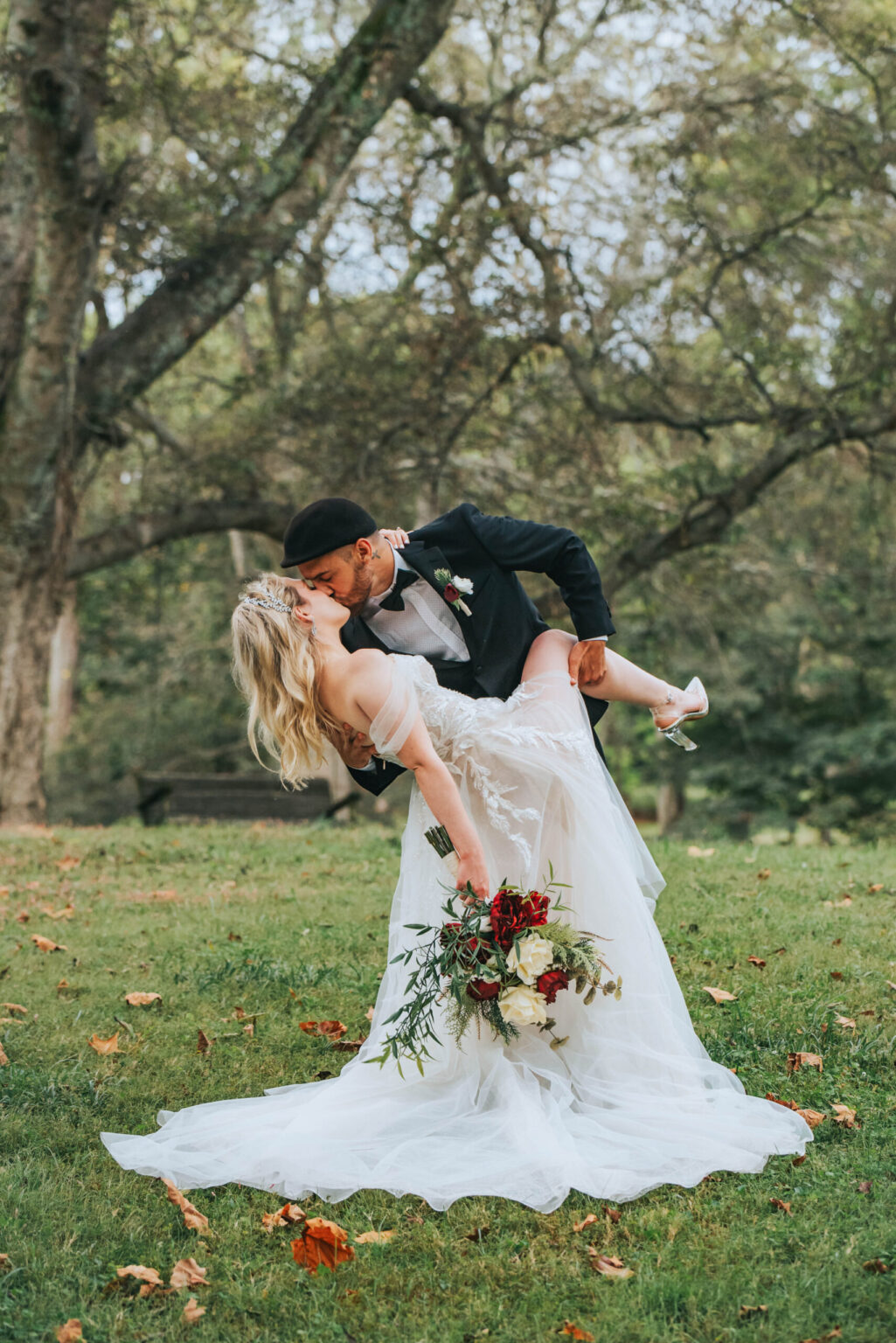 Elopement Wedding A bride and groom share a romantic kiss outdoors under a large, leafy tree. The groom, wearing a dark suit, gently dips the bride, who wears a flowing white gown and holds a vibrant bouquet of red and white flowers. The grassy ground is dotted with fallen autumn leaves. Elopements Inc