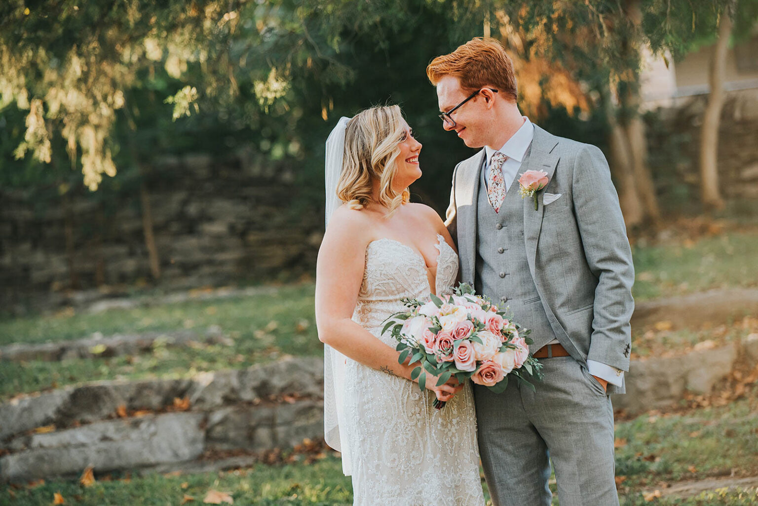 Elopement Wedding A bride and groom stand outdoors, smiling at each other. The bride wears a white lace wedding dress and holds a bouquet of white and pink flowers. The groom, with red hair and glasses, wears a light gray suit with a white shirt, floral tie, and a pink boutonniere. Trees and stone steps are in the background. Elopements Inc