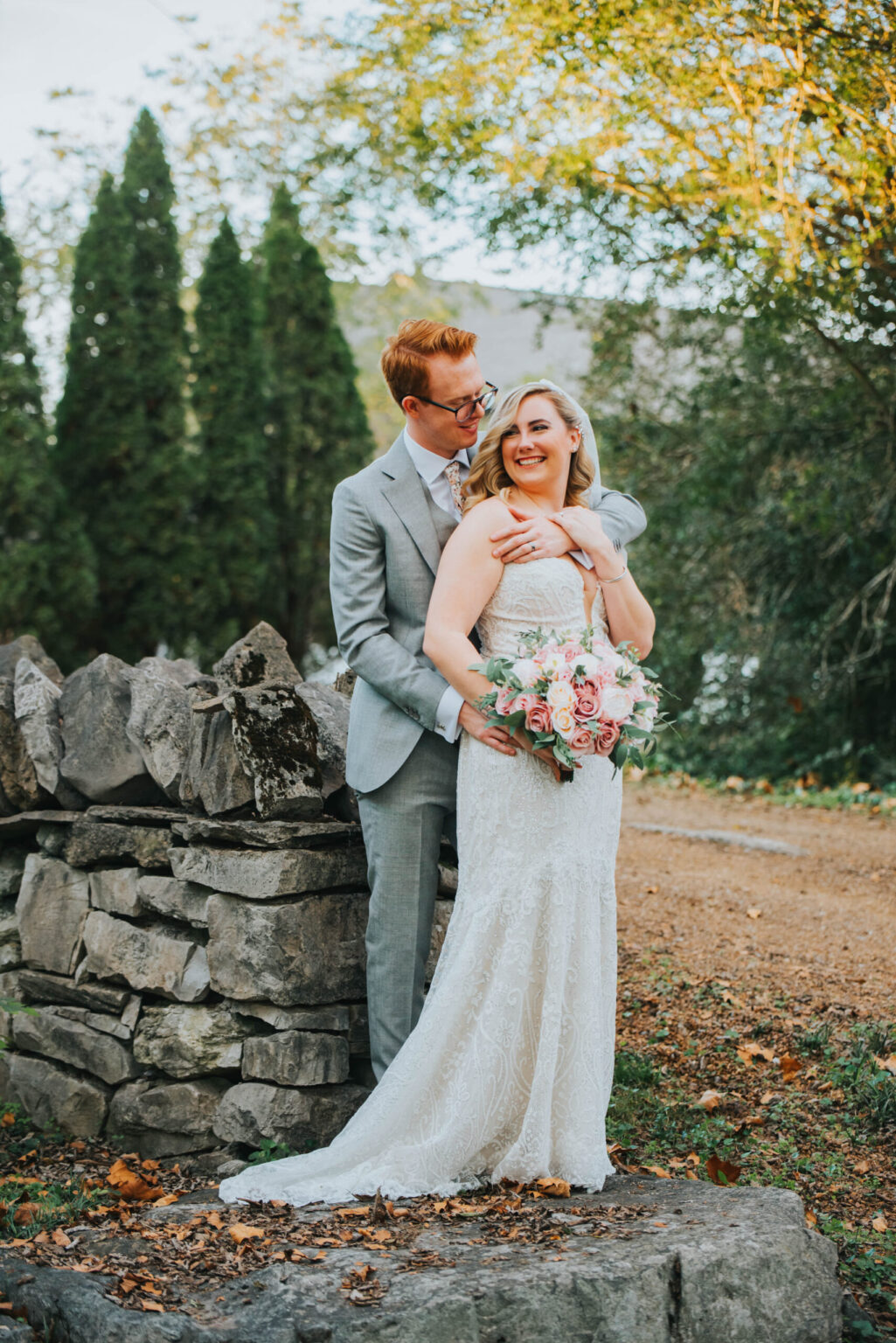 Elopement Wedding A bride and groom embrace in an outdoor setting, standing by a stone wall. The bride wears a strapless white lace gown and holds a bouquet of pink and white flowers. The groom, wearing a light gray suit, stands behind her, smiling. The background features trees and a pathway covered with fallen leaves. Elopements Inc