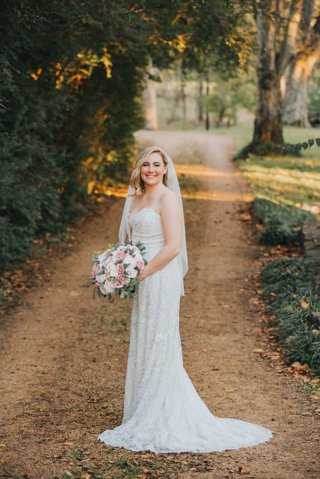 Elopement Wedding A smiling bride in a strapless white lace wedding gown and veil holds a bouquet of pink and white flowers. She stands on a sunlit dirt path surrounded by lush greenery and trees, looking towards the camera. It appears to be late afternoon with soft, warm sunlight illuminating the scene. Elopements Inc