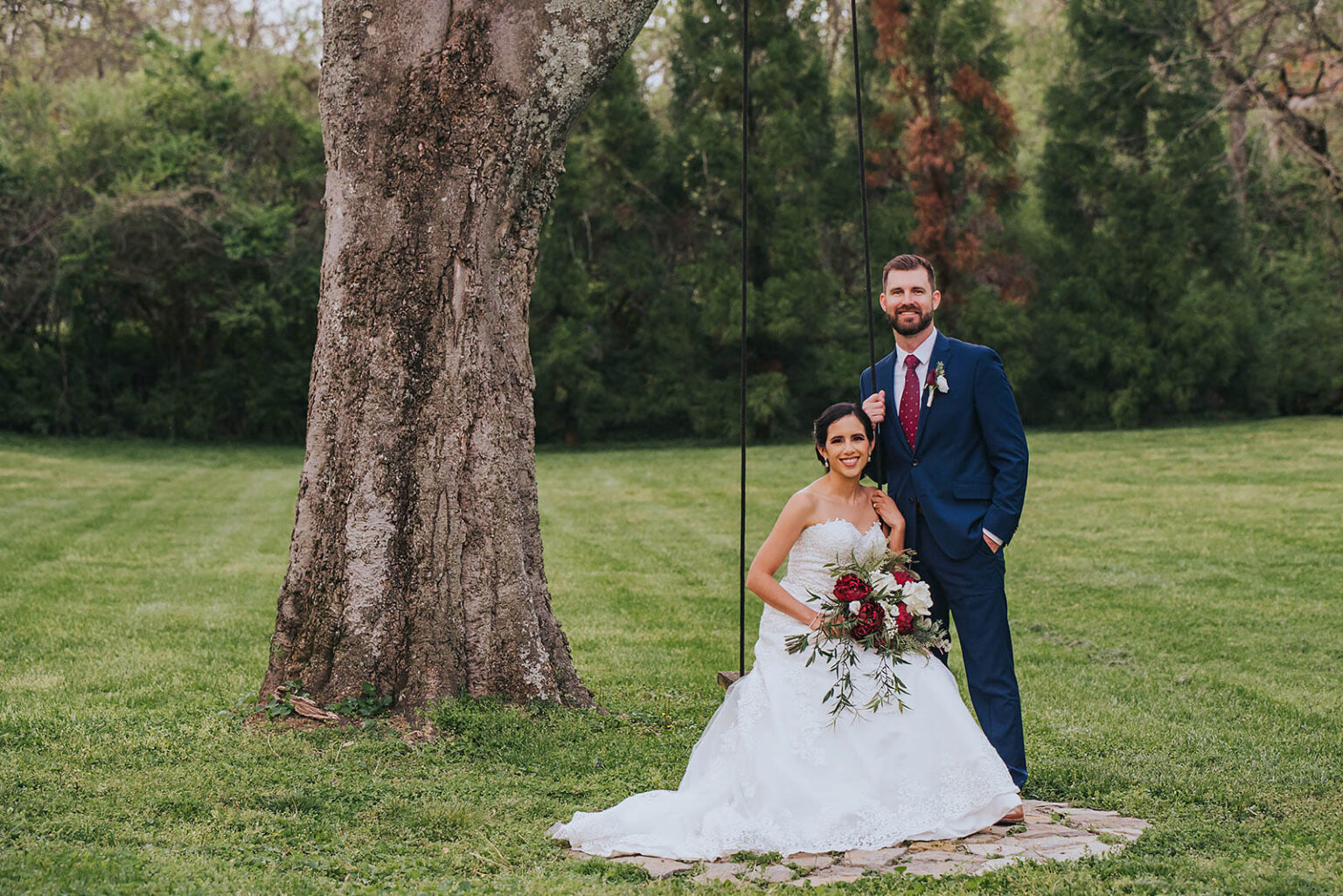 Elopement Wedding A bride and groom pose together for a wedding photo outdoors. The bride is seated in front, wearing a white gown and holding a bouquet of red and white flowers. The groom stands behind her, wearing a navy suit with a red tie. They are under a large tree in a green, grassy area with forested background. Elopements Inc