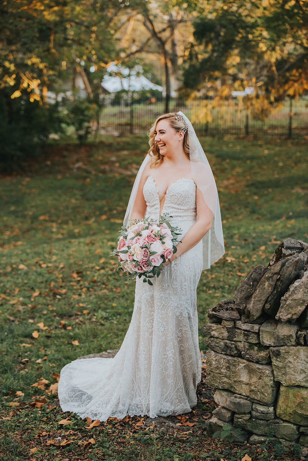 Elopement Wedding A bride wearing a lace wedding gown is standing outside on a grassy area, smiling. She is holding a bouquet of pink and white flowers. A veil drapes over her shoulders, and trees with fall foliage form the background. She stands near a short stone wall. Elopements Inc