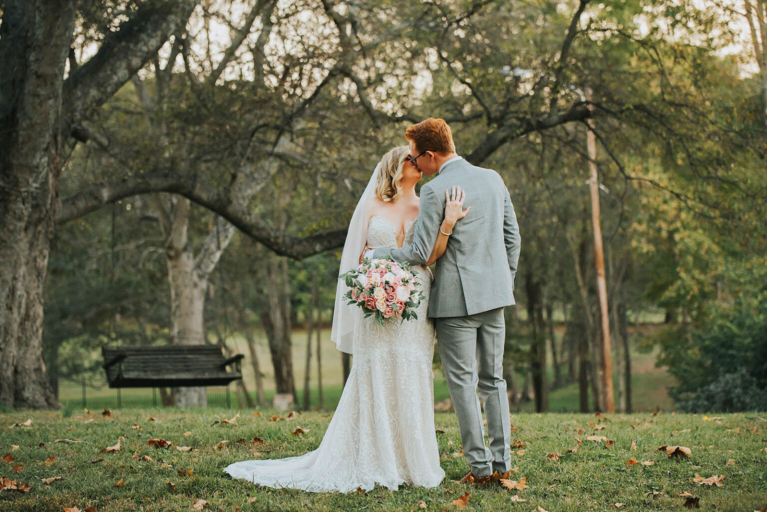 Elopement Wedding A bride in a white lace gown and veil kisses a groom in a light gray suit outdoors. The bride holds a bouquet of pink and white flowers. They stand on a grassy area scattered with fallen leaves, with large trees and a bench in the background. The scene is serene and filled with natural light. Elopements Inc