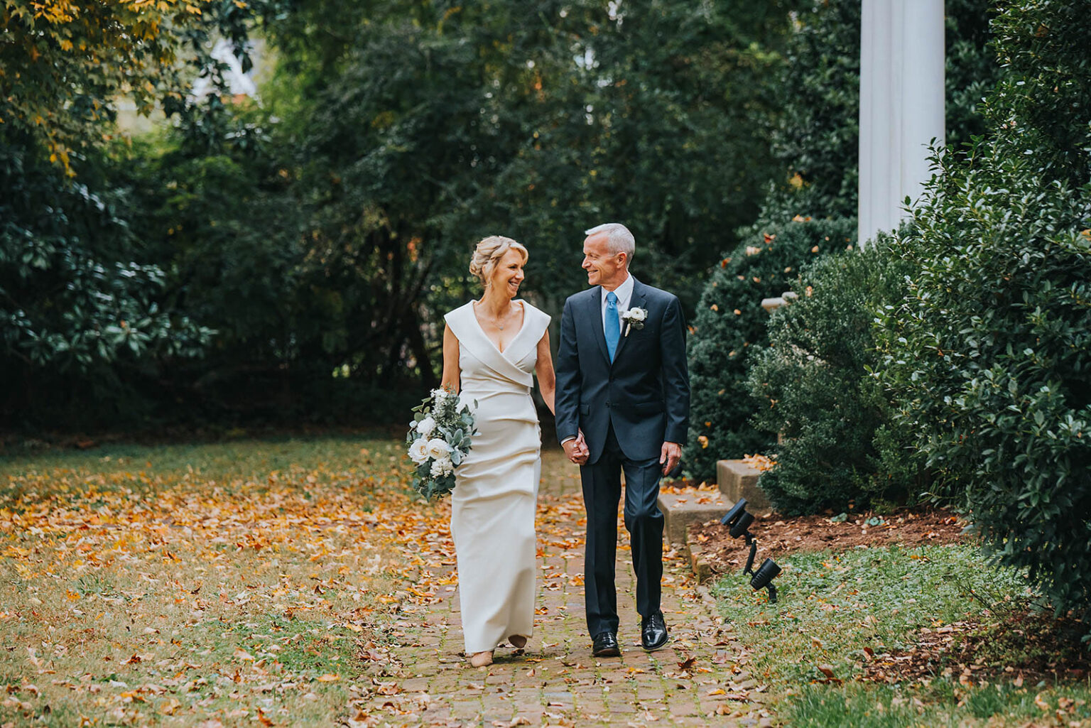 Elopement Wedding A bride in a white dress and a groom in a dark suit walk hand in hand down a brick pathway. They are surrounded by lush greenery and foliage, with autumn leaves scattered on the ground. Both are smiling at each other warmly. The background features trees and a white column. Elopements Inc