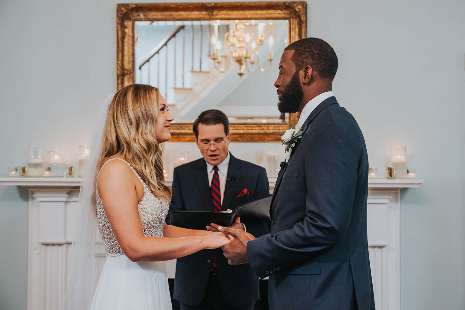 Elopement Wedding A couple stands facing each other, holding hands, during a wedding ceremony. The bride, in a white dress with a beaded top, and the groom, in a navy suit, are smiling. An officiant, holding a book, stands behind them in front of a fireplace. A large mirror and candles adorn the background. Elopements Inc