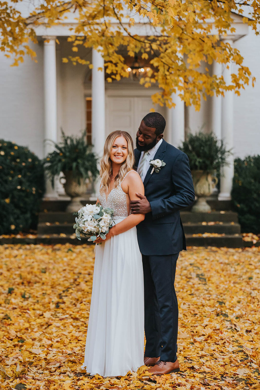 Elopement Wedding A couple stands closely, smiling, surrounded by yellow autumn leaves. The bride, in a white gown, holds a bouquet of white flowers and greenery. The groom, in a navy blue suit, stands behind her with hands gently on her waist. They are in front of a white building with large columns and greenery. Elopements Inc