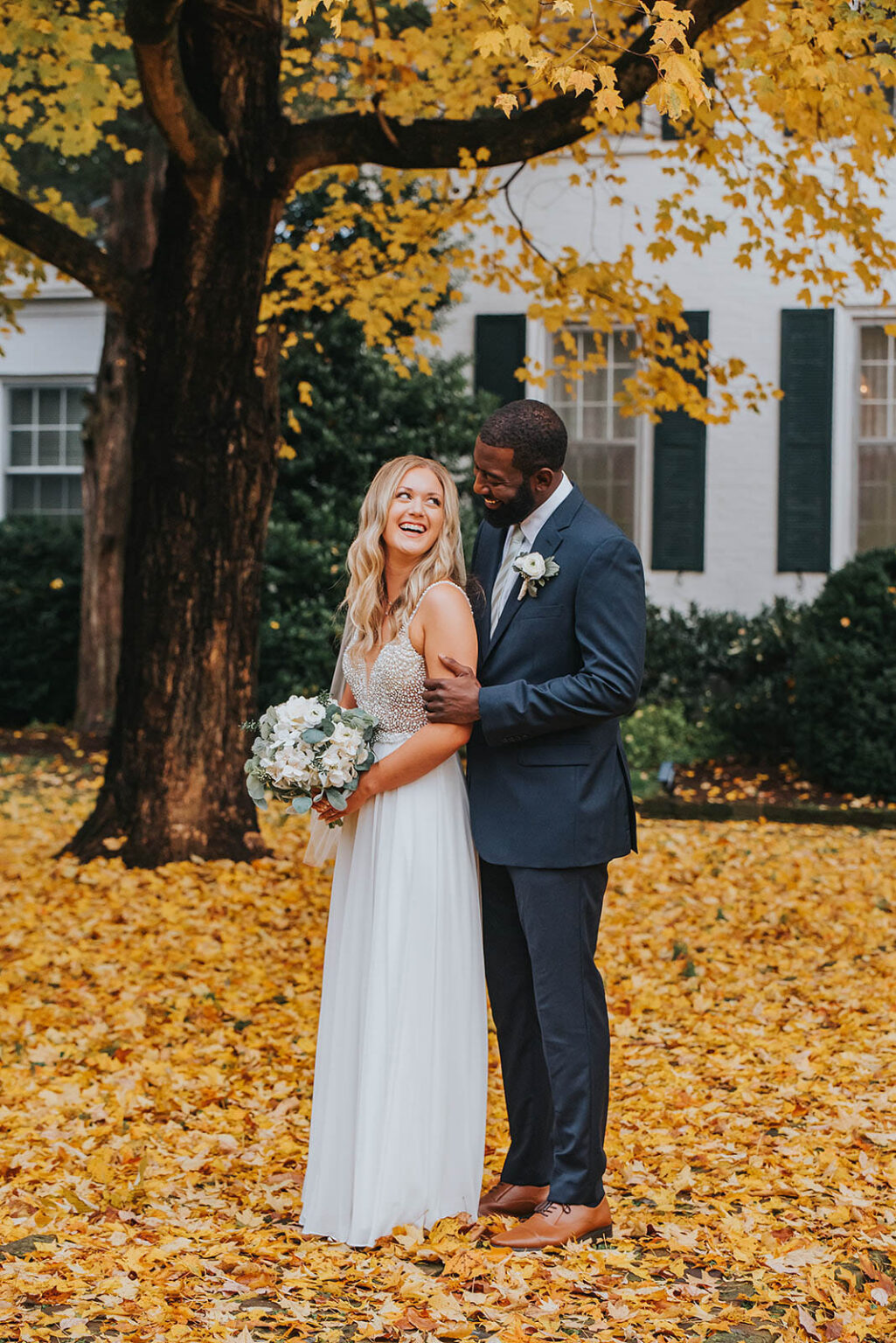 Elopement Wedding A bride and groom are standing under a large tree with yellow autumn leaves covering the ground. The bride, in a white dress and holding a bouquet, smiles joyfully at the groom in a dark suit who stands behind her, embracing her with a loving expression. A white building with green shutters is in the background. Elopements Inc