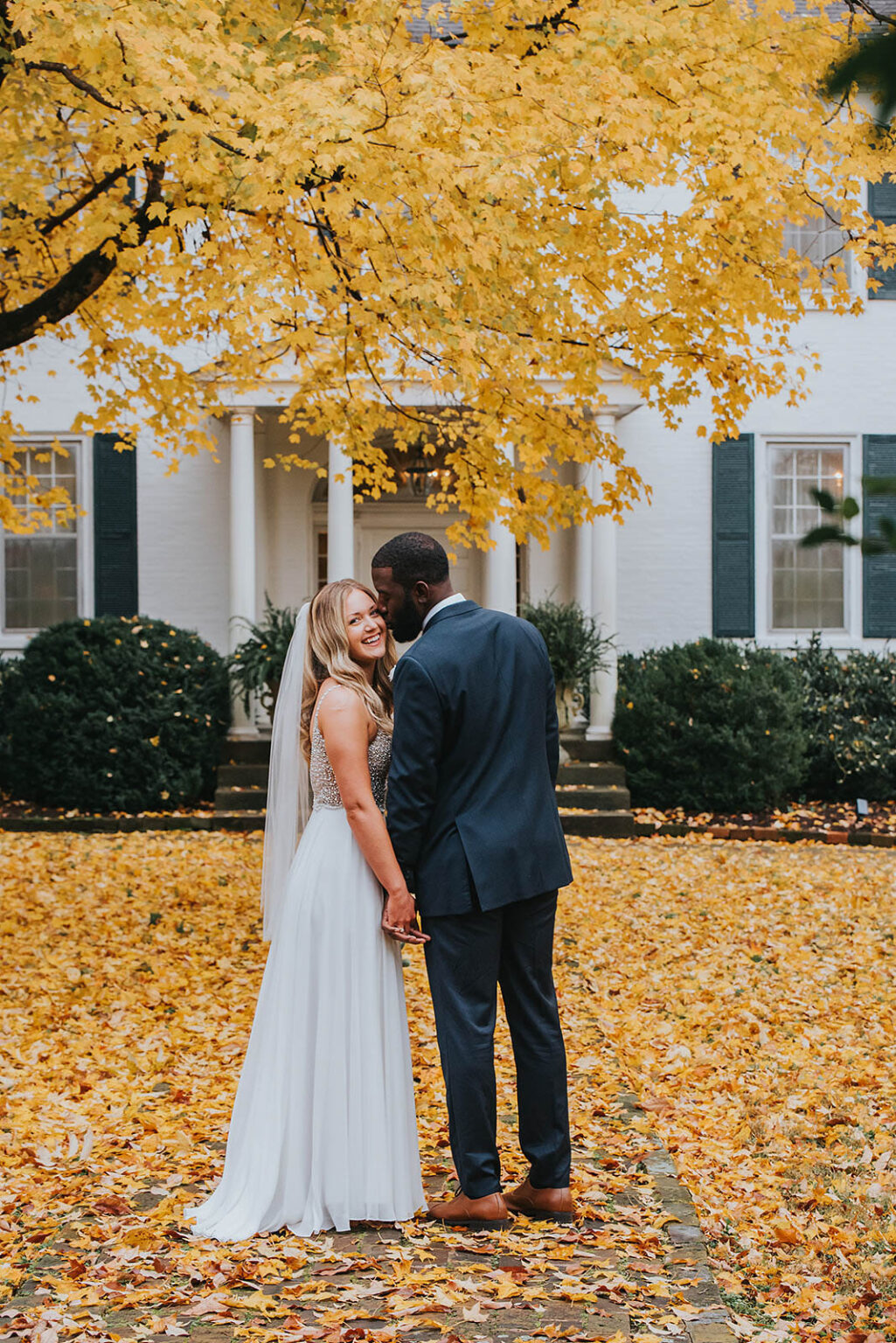 Elopement Wedding A bride and groom stand close, holding hands and facing each other under a tree with vibrant yellow leaves. The bride wears a flowing white gown and a veil, while the groom is in a dark suit. They are at an outdoor location with a white colonial-style house in the background surrounded by fallen leaves. Elopements Inc