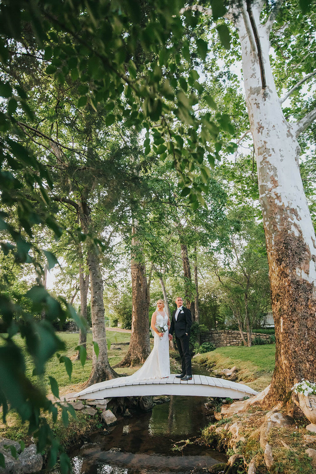 Elopement Wedding A bride in a white gown and a groom in a black suit stand hand in hand on a small wooden bridge over a creek in a lush, green forested area. The scene is framed by tree branches and leaves, creating a natural, serene setting as sunlight filters through the canopy. Elopements Inc