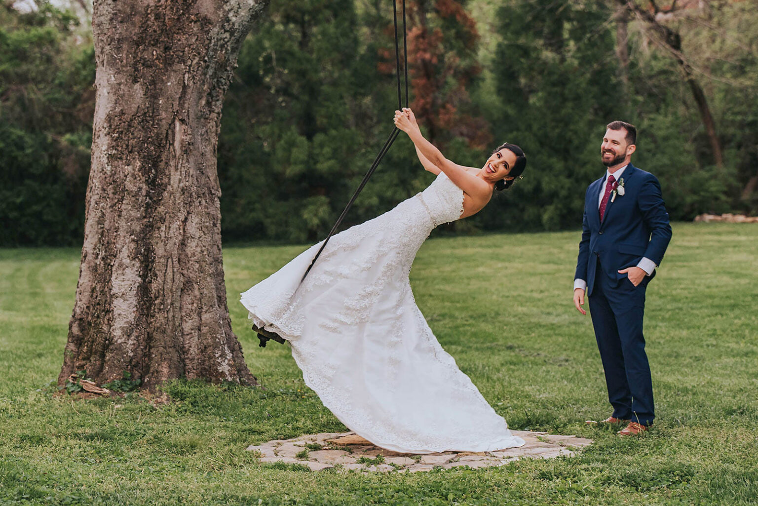 Elopement Wedding A bride in a white wedding gown joyfully swings on a rope swing attached to a large tree at an outdoor setting, smiling widely. The groom, dressed in a dark blue suit with a red tie, stands nearby, looking at her with a smile. The background is filled with lush greenery. Elopements Inc