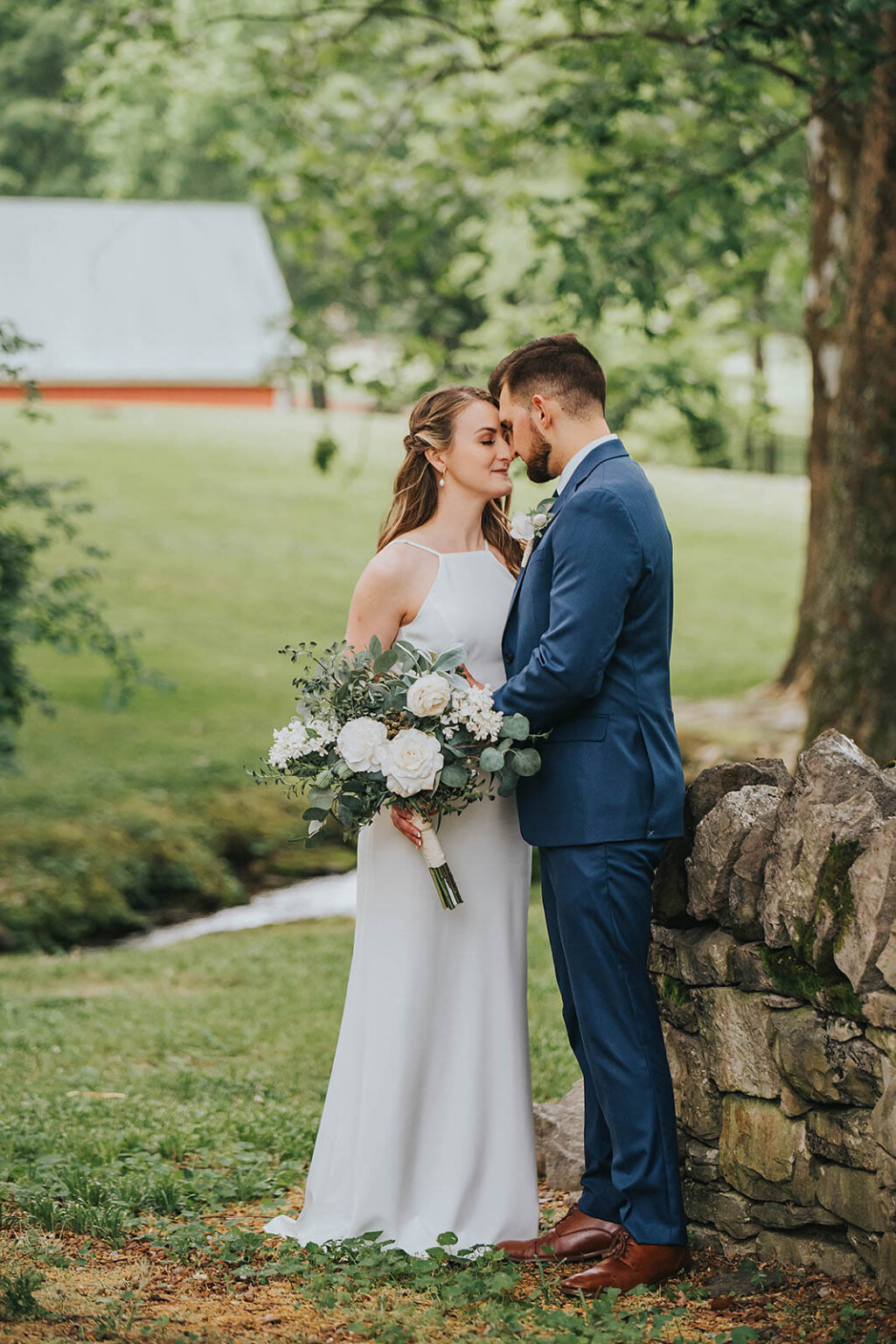 Elopement Wedding A bride in a white dress and a groom in a blue suit share an intimate moment outdoors. They are standing closely, touching foreheads, next to a stone wall. The bride holds a bouquet of white and pastel flowers. A green lawn, trees, and a white barn with a red foundation are visible in the background. Elopements Inc