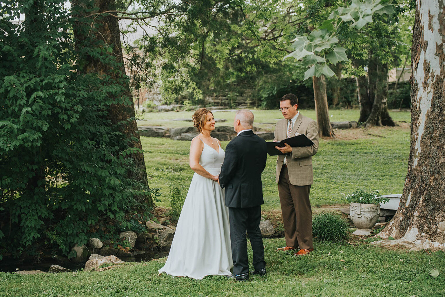 Elopement Wedding A couple is getting married outdoors. The bride, in a white dress, and the groom, in a black suit, are facing each other holding hands. An officiant, dressed in a brown jacket and orange shoes, stands behind them with an open book. Trees and grass surround them, creating a serene backdrop. Elopements Inc