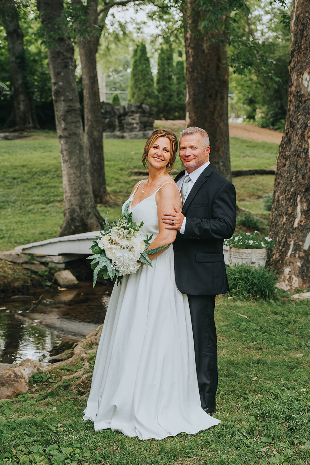 Elopement Wedding A bride and groom stand outdoors, dressed in wedding attire. The bride, in a white gown, holds a bouquet of white and green flowers. The groom, in a dark suit with a light tie, stands behind her. They pose in a lush garden setting with tall trees and a small stream in the background. Elopements Inc