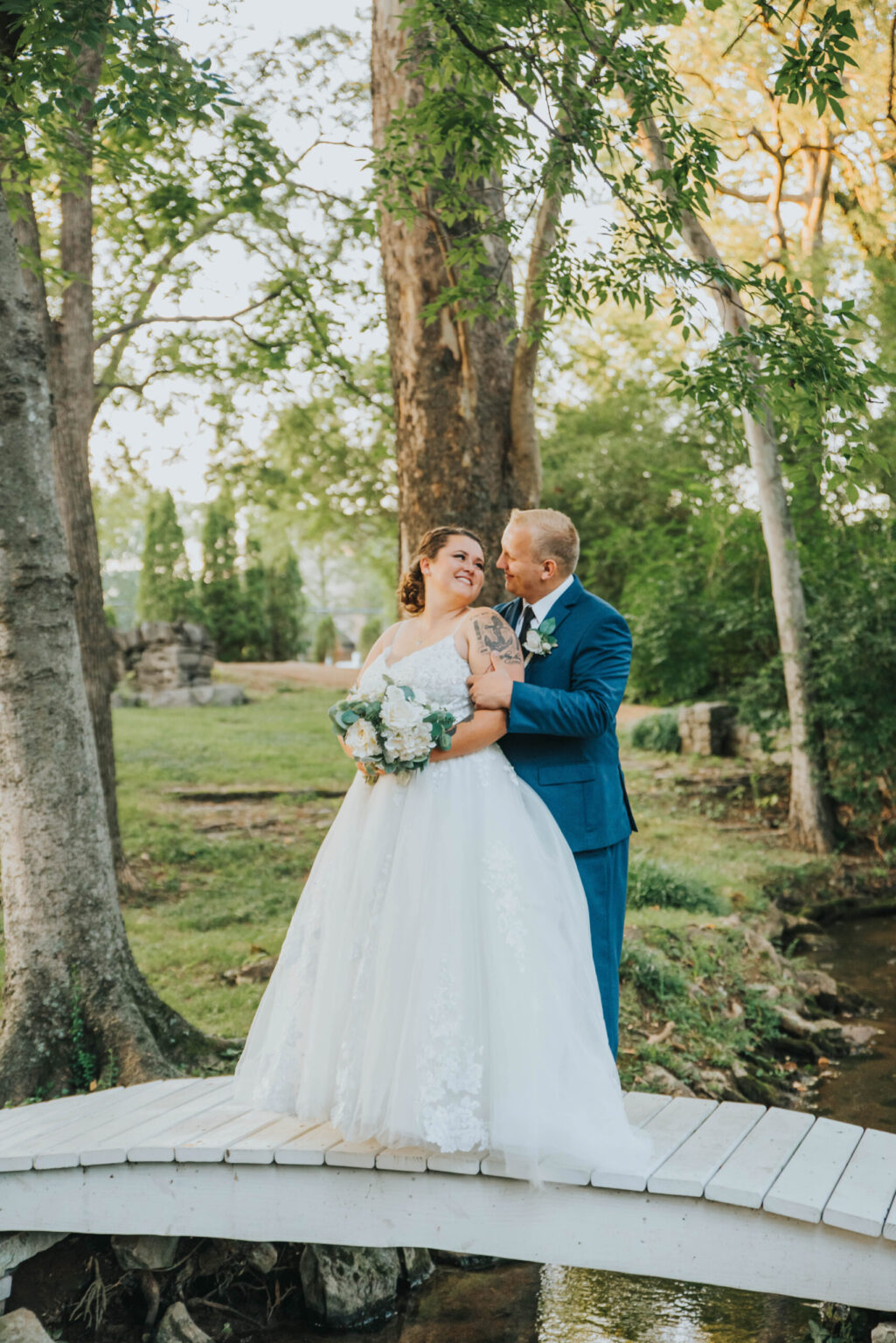 Elopement Wedding A bride and groom stand on a small white bridge in a serene wooded area. The bride wears a white gown and holds a bouquet, while the groom, in a blue suit, embraces her from behind, both smiling. The background includes trees, lush greenery, and a small creek flowing underneath the bridge. Elopements Inc