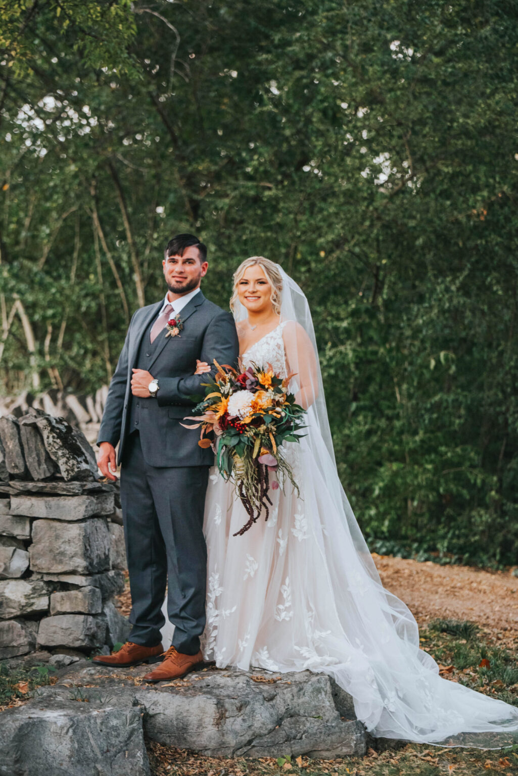 Elopement Wedding A bride and groom stand outdoors, surrounded by greenery. The groom is in a gray suit with a mauve tie, and the bride wears a white, lace wedding gown with a long veil. She holds a bouquet featuring various flowers and greenery. Behind them is a stone wall and a tree-lined backdrop. Elopements Inc