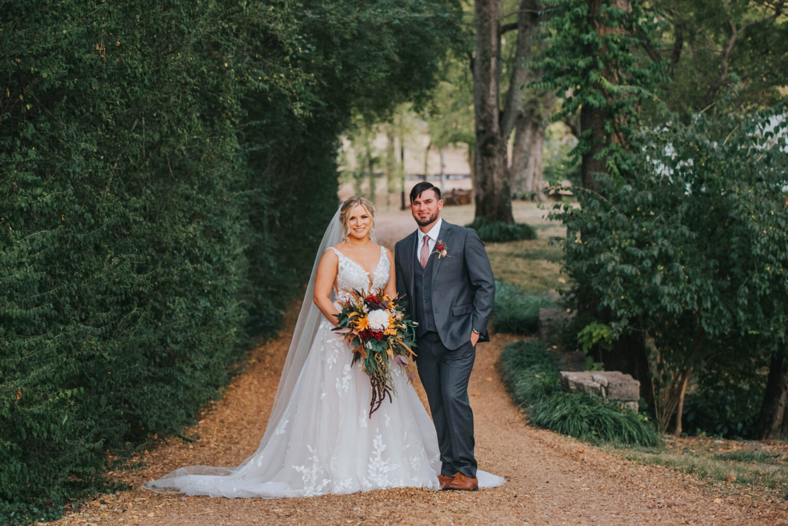 Elopement Wedding A bride and groom stand on a gravel path flanked by lush greenery. The bride wears a white lace dress and veil, holding a vibrant bouquet of flowers. The groom is in a dark suit with a pink tie and boutonnière. Both smile and face the camera, surrounded by a peaceful, tree-filled outdoor setting. Elopements Inc