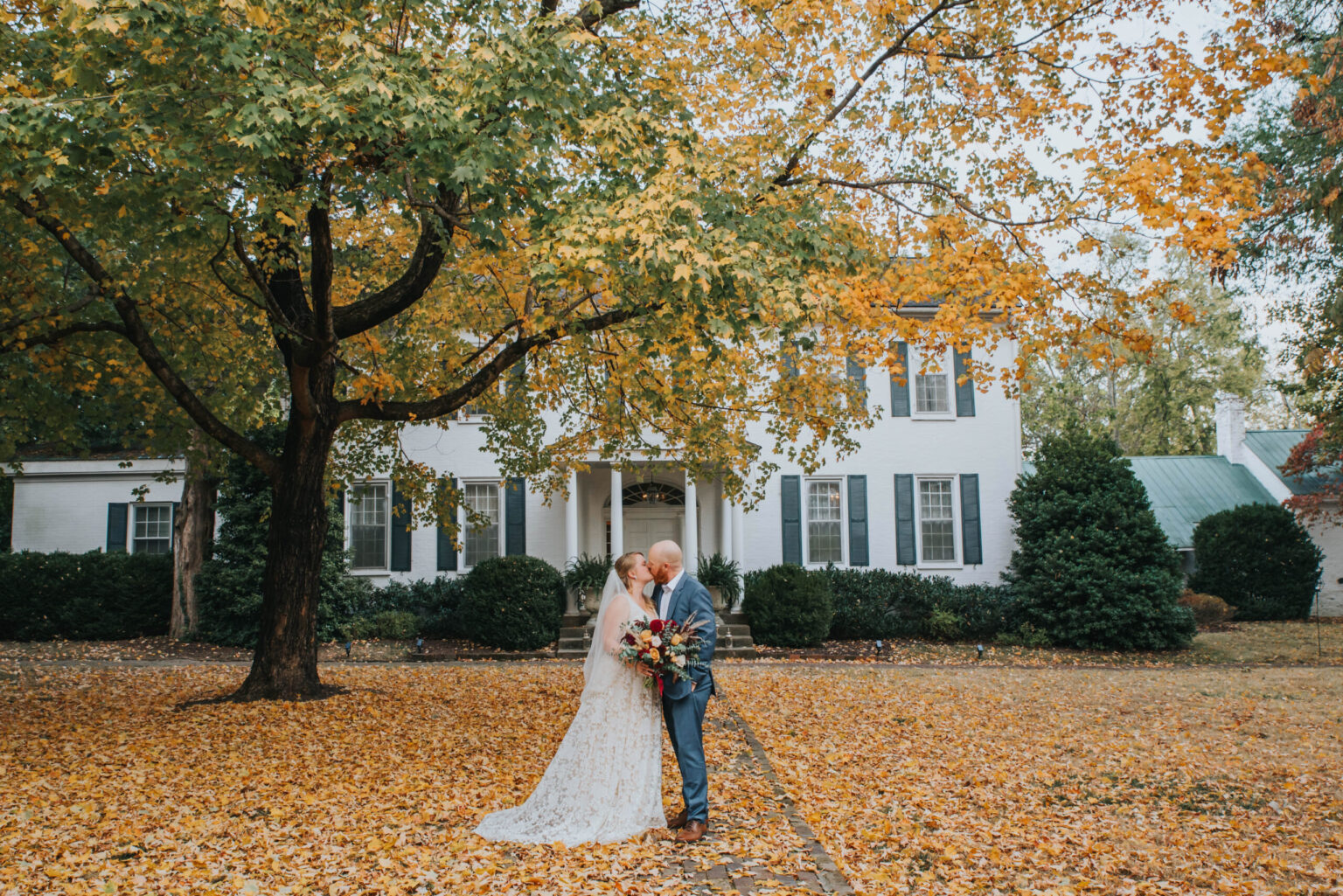 Elopement Wedding A bride and groom share a kiss under a large tree with golden autumn leaves in front of a white colonial-style house. The bride is in a white lace gown, holding a bouquet, while the groom is in a blue suit. The yard is covered in fallen leaves, creating a picturesque fall scene. Elopements Inc