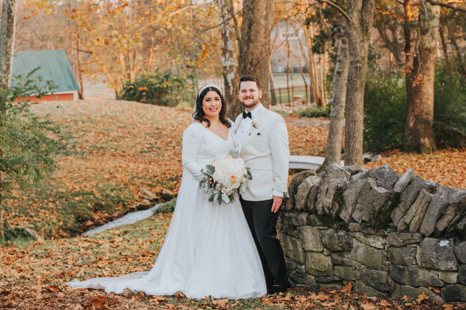 Elopement Wedding A bride and groom stand in a rustic outdoor setting with autumn foliage. The bride wears a white dress and holds a bouquet of pink and white flowers. The groom is in a white tuxedo jacket and black pants. They are leaning against a stone wall, smiling at the camera. Elopements Inc