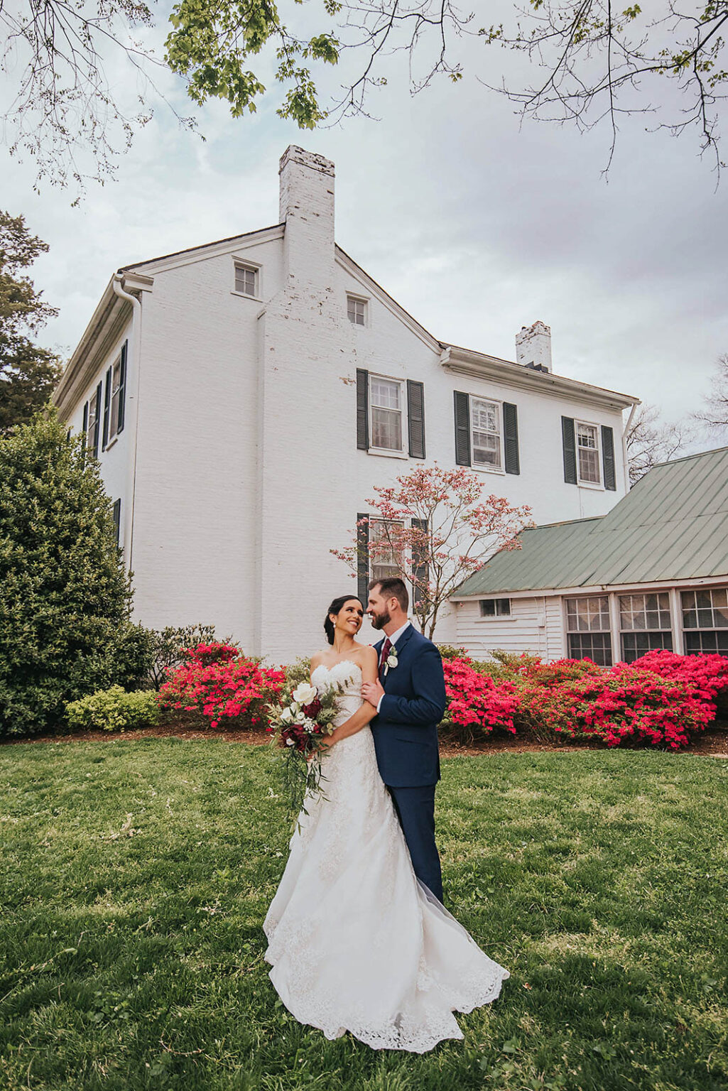 Elopement Wedding A bride in a white lace gown and a groom in a navy suit stand closely together, holding a bouquet of flowers. They are in front of a large white house with black shutters. The yard is lush with green grass, vibrant pink and red azaleas, and trees with budding leaves in the background. Elopements Inc