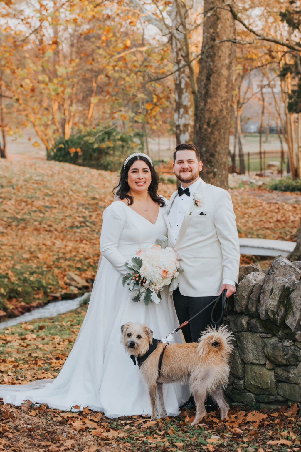 Elopement Wedding A newlywed couple poses outdoors in early autumn. The bride, in a white gown with long sleeves, holds a bouquet of white and peach flowers. The groom wears a white jacket and black bow tie. They stand near a stone wall, with a small beige dog on a leash in front of them. Fallen leaves cover the ground. Elopements Inc