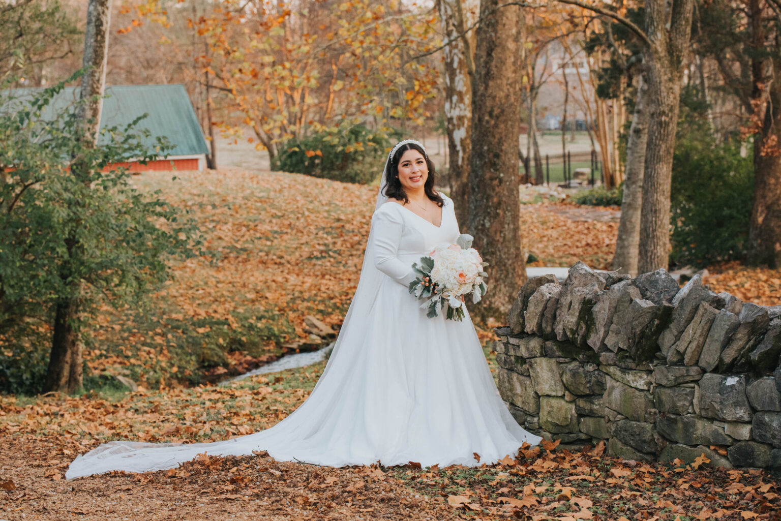 Elopement Wedding A bride in a white wedding gown stands outside near a stone wall, holding a bouquet of white flowers. The background features an autumnal scene with fallen leaves, trees, and a distant green-roofed building. The bride has a gentle smile and the train of her dress flows behind her. Elopements Inc