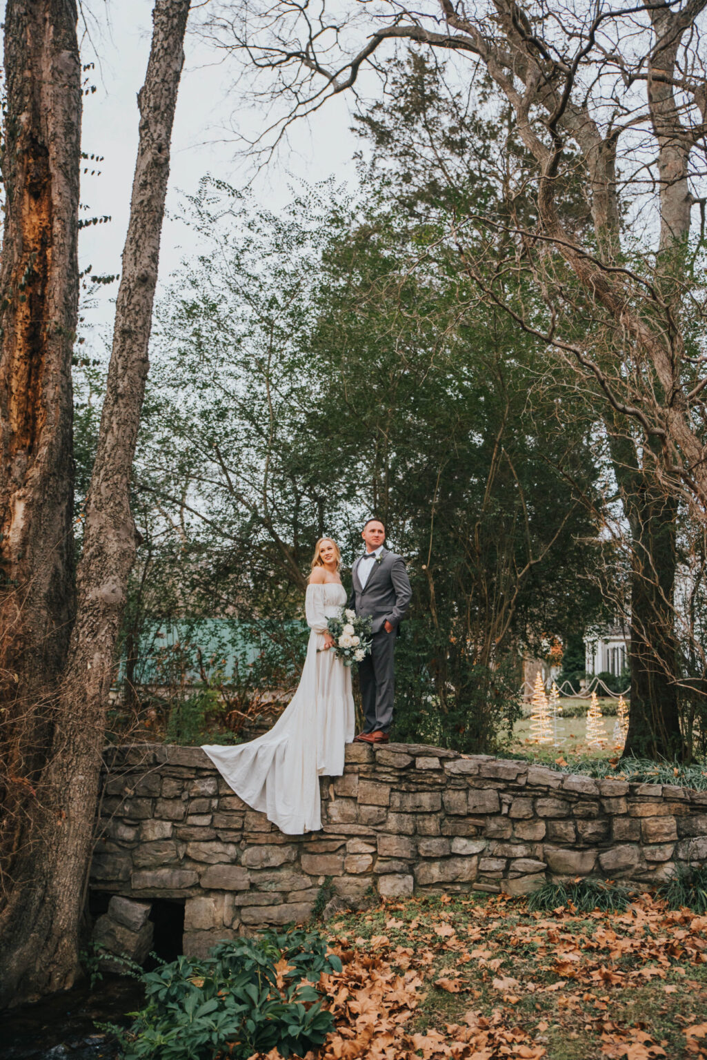 Elopement Wedding A bride in a white dress and a groom in a gray suit stand on a stone bridge surrounded by trees and foliage. The bride holds a bouquet of white flowers. The scene is outdoors, and the background features bare trees and some greenery, indicating a late autumn or early winter setting. Elopements Inc