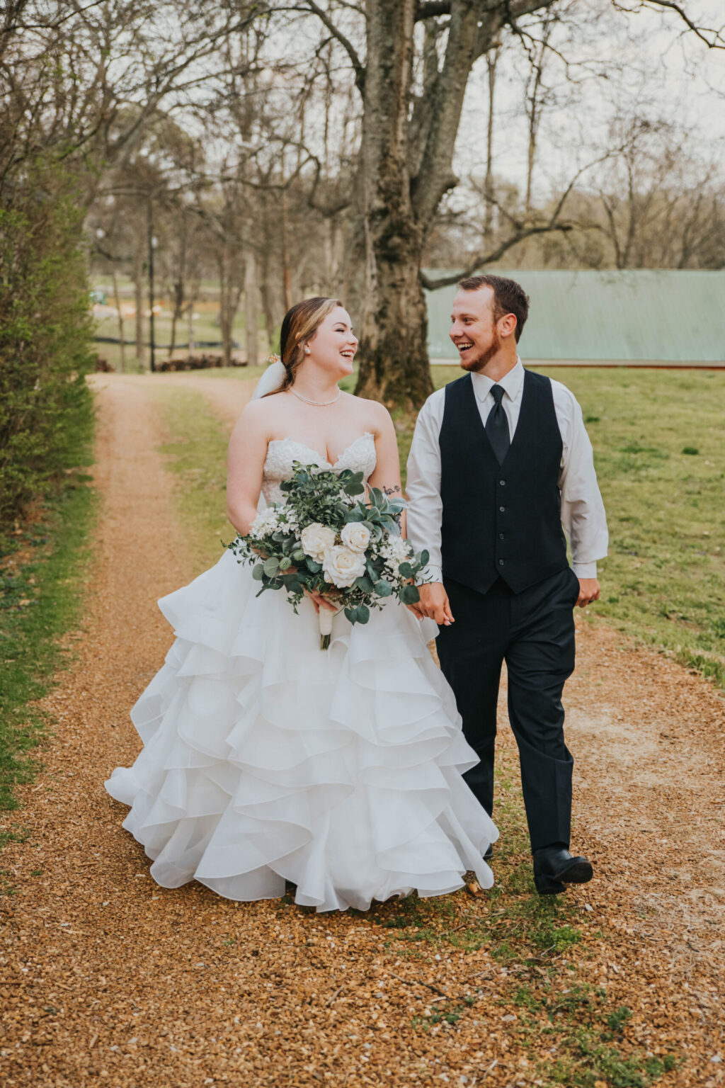 Elopement Wedding A smiling bride and groom walk hand-in-hand down a tree-lined gravel path. The bride wears a white, layered gown and holds a bouquet of roses and greenery. The groom is dressed in a dark vest and tie over a white shirt. The background features trees, grass, and a distant green-roofed structure. Elopements Inc