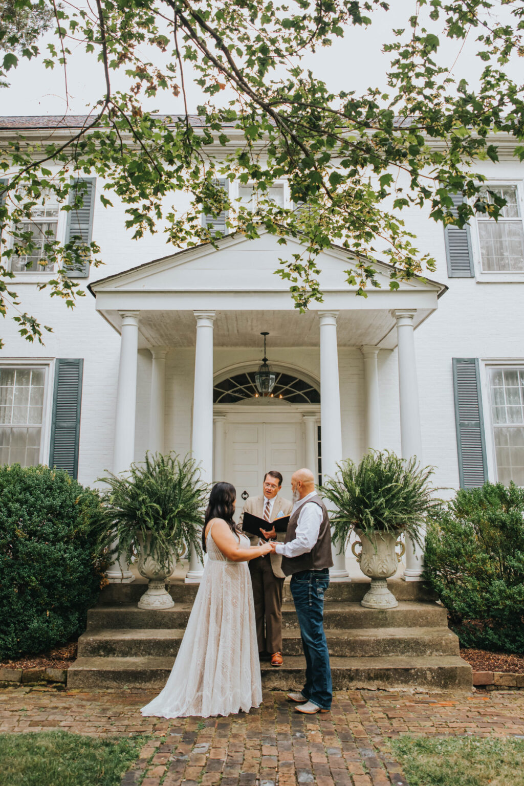 Elopement Wedding A couple is getting married in front of a large white colonial-style house with green shutters. The bride in a white dress and the groom in jeans and a vest hold hands while facing the officiant. They stand on a stone step pathway, framed by lush greenery and ferns, under an overcast sky. Elopements Inc