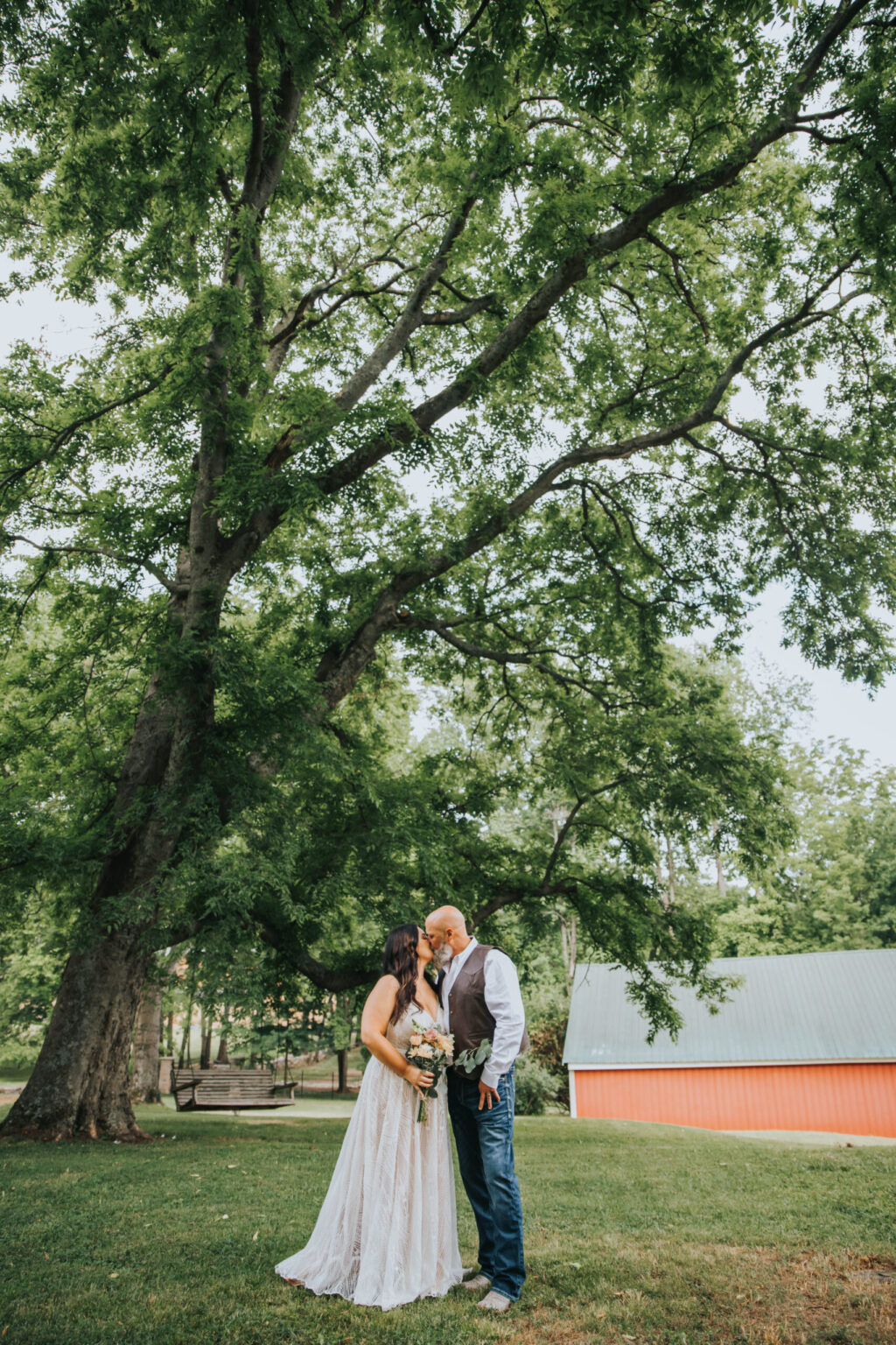 Elopement Wedding A couple stands under a large, leafy tree in a serene outdoor setting. The woman, in a white gown, holds a bouquet and gazes at the man, who is dressed in a vest and jeans. Behind them is a red barn with a grey roof. They stand close, touching foreheads, creating an intimate moment. Elopements Inc