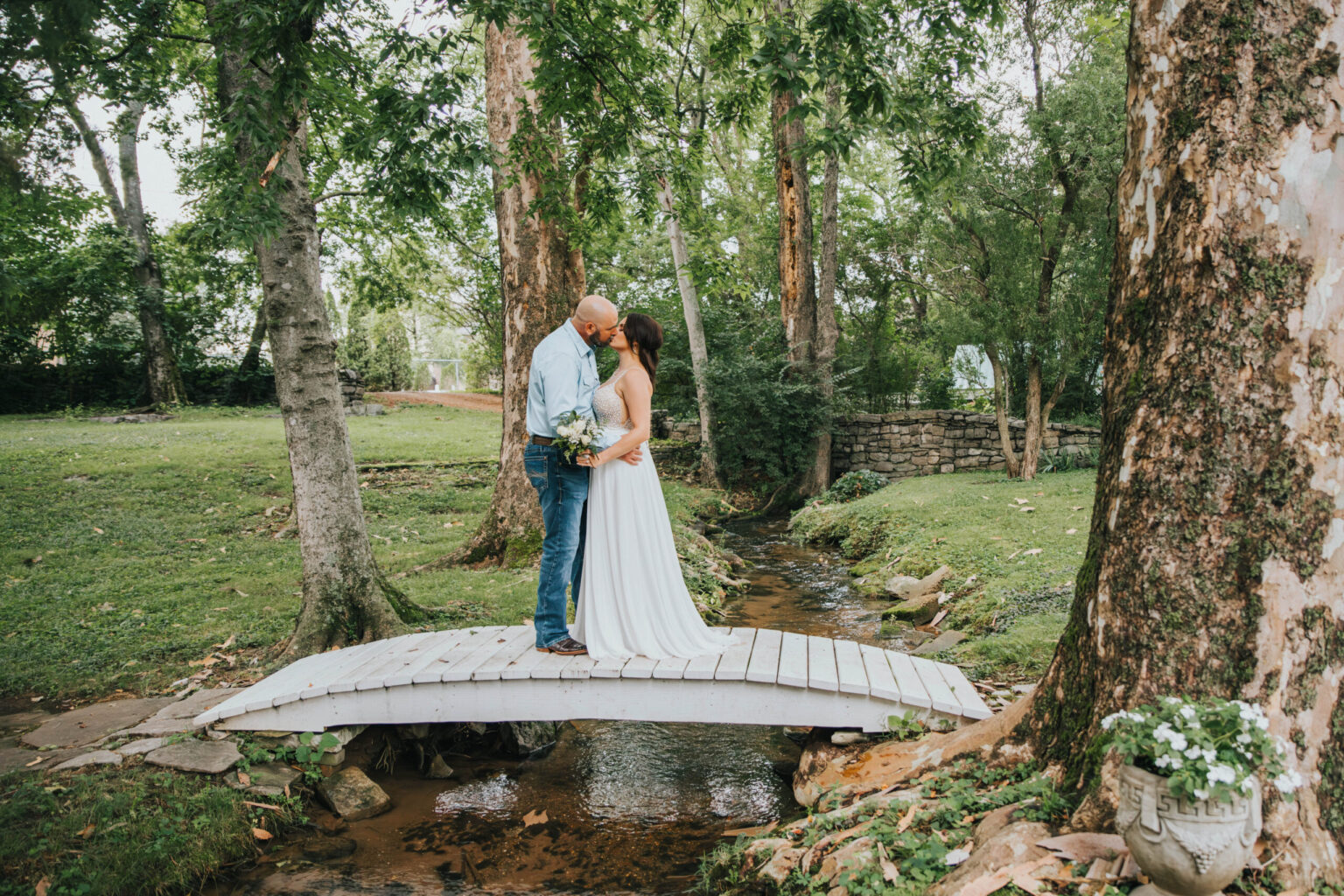 Elopement Wedding A couple embraces and kisses on a small, white wooden bridge over a stream in a lush garden. The woman, wearing a white wedding dress and holding a bouquet, stands with the man, who is dressed in jeans and a light blue shirt. Surrounding them are tall trees and green grass, with decorative stone planters. Elopements Inc