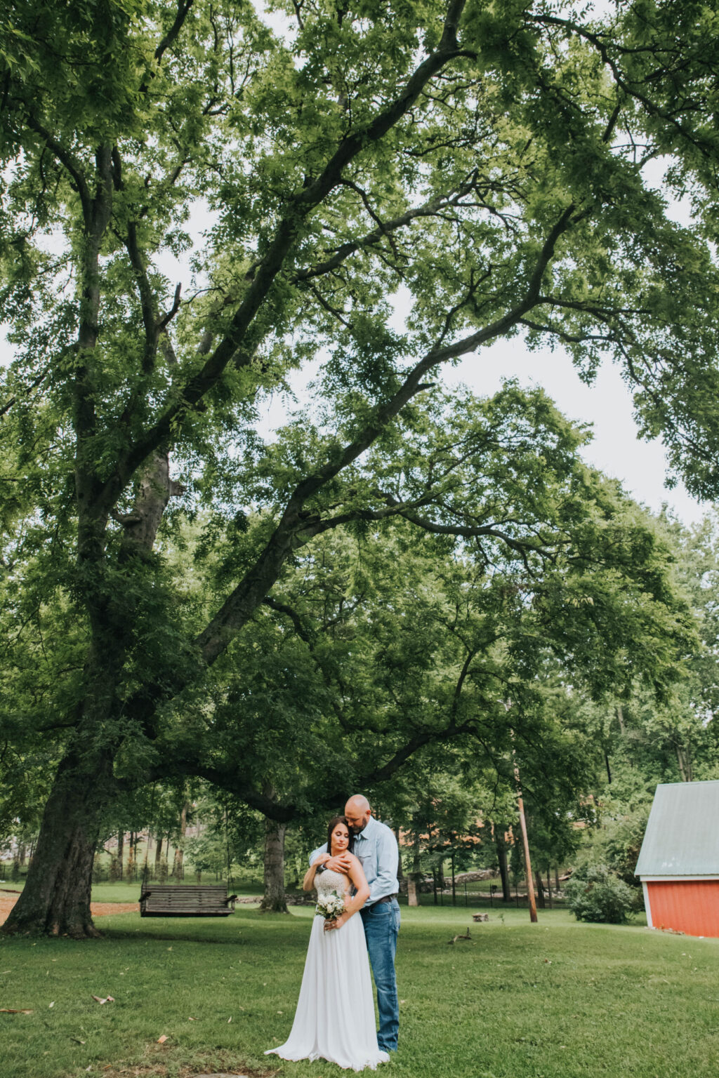 Elopement Wedding A couple stands in an open grassy area under a large, lush tree. The woman, dressed in a white gown, holds a bouquet and smiles while the man, wearing a blue shirt and jeans, embraces her from behind. In the background, there is a red barn and a wooden bench swing hanging from another tree. Elopements Inc