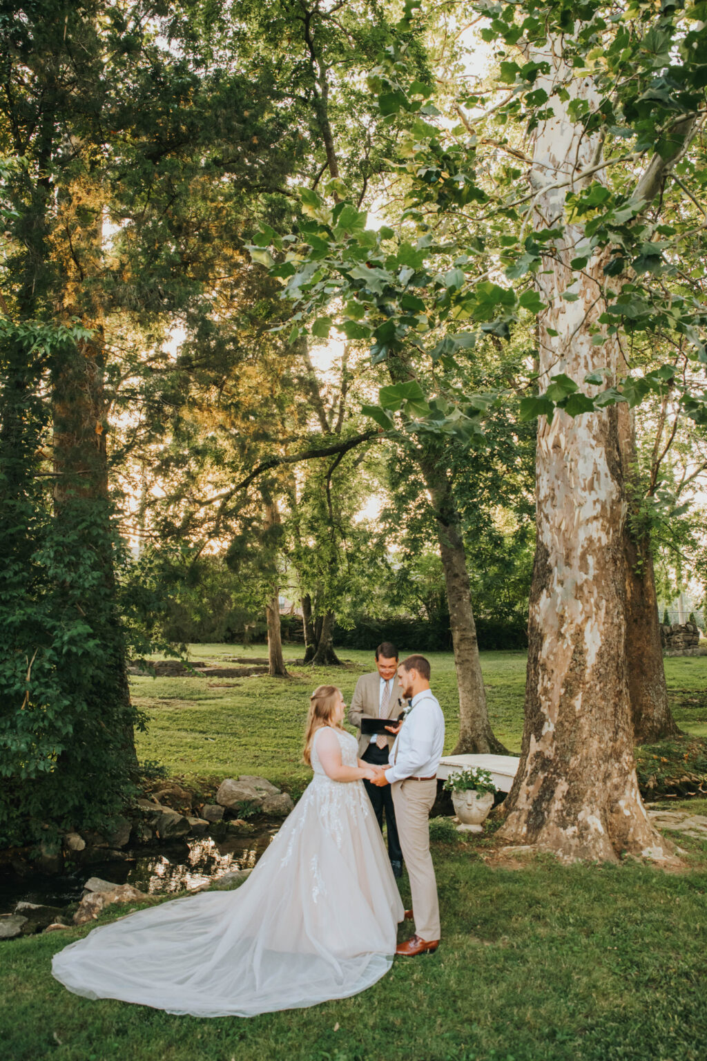 Elopement Wedding A couple stands outdoors exchanging vows in front of an officiant. The bride wears a long, flowing white gown, and the groom is in a white shirt and light-colored trousers. They are surrounded by lush greenery, a large tree, and a stream, creating a serene and picturesque setting for the ceremony. Elopements Inc