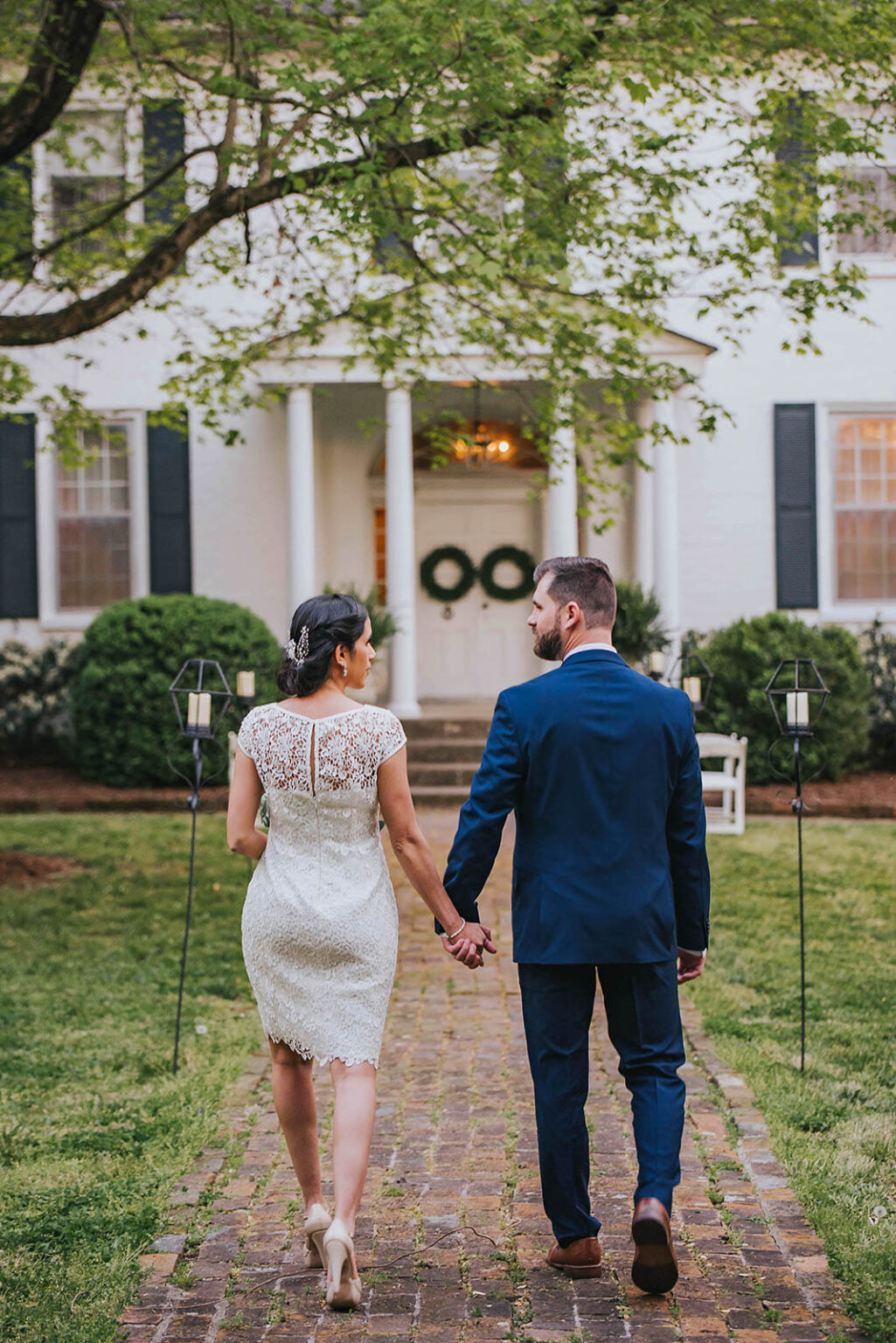 Elopement Wedding A couple dressed in formal attire walks hand in hand towards a white building adorned with wreaths on its doors. The woman is in a lace white dress and heels, and the man is in a navy blue suit and brown shoes. They are on a brick path surrounded by greenery and lanterns along the sides. Elopements Inc