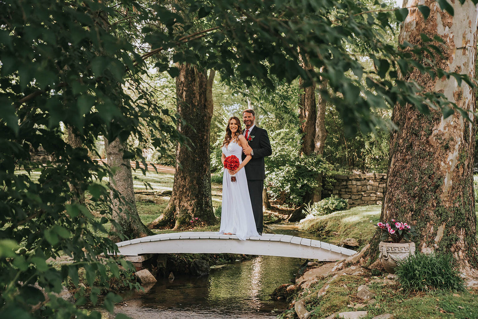 Elopement Wedding A bride and groom stand closely together on a small white bridge over a creek in a lush, green garden. The bride holds a bouquet of bright red flowers, and the groom wears a dark suit. Trees and foliage frame the scene, adding a tranquil, nature-rich backdrop to the couple's moment. Elopements Inc