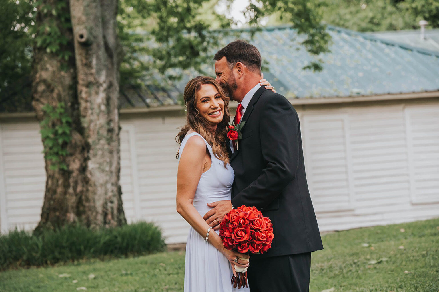 Elopement Wedding A smiling couple stands close in an outdoor setting, dressed in formal attire. The woman in a light blue gown holds a red rose bouquet, and the man in a black suit with a red boutonniere leans in affectionately. They are in front of a building with a green roof, surrounded by greenery and trees. Elopements Inc