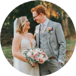 Elopement Wedding A bride and groom share a joyful gaze on their wedding day. The bride, in a white lace dress and veil, holds a bouquet of pink and white roses. The groom, in a gray suit, white shirt, and floral tie, wears glasses. They stand in an outdoor setting with green foliage in the background. Elopements Inc